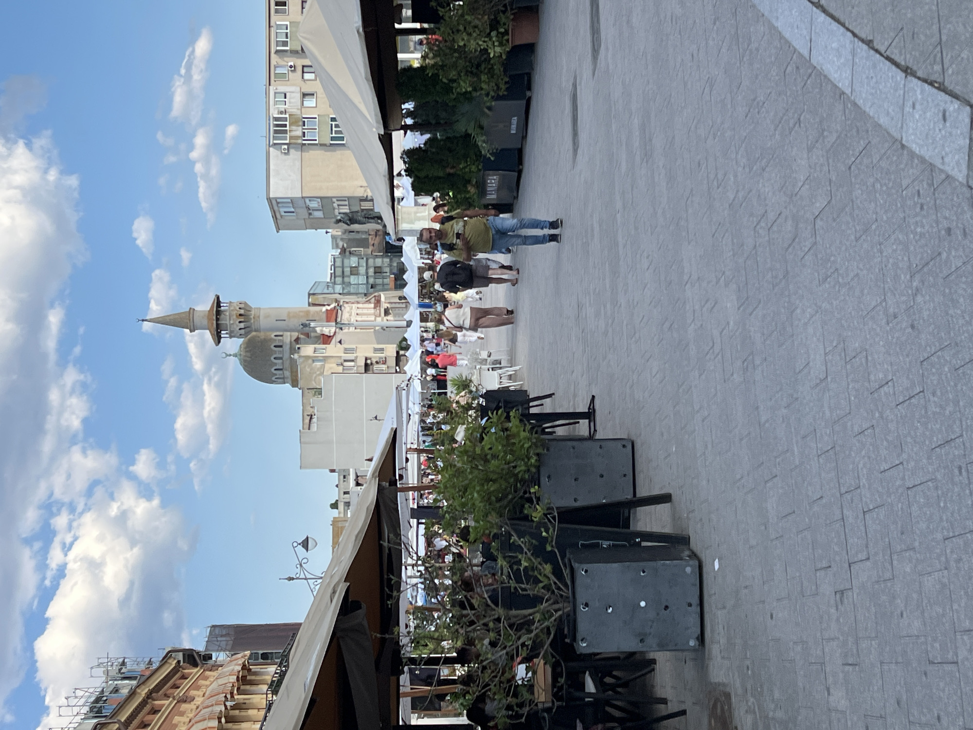 This is a ground-level, full-shot, outdoor photograph taken on a paved pedestrian area in Constanța, Romania. The scene depicts a street with outdoor seating for businesses, characterized by large umbrellas providing shade over tables and chairs. In the foreground, there are planters with lush green foliage. Several people are walking along the street. In the mid-ground, there are white tents set up, suggesting a market or outdoor event. Dominating the background skyline is a mosque with a distinctive dome and minaret, an architectural element characteristic of the city's historical influences. The sky above is partly cloudy, with a clear blue expanse. The overall impression is of a lively public space on a bright day.
