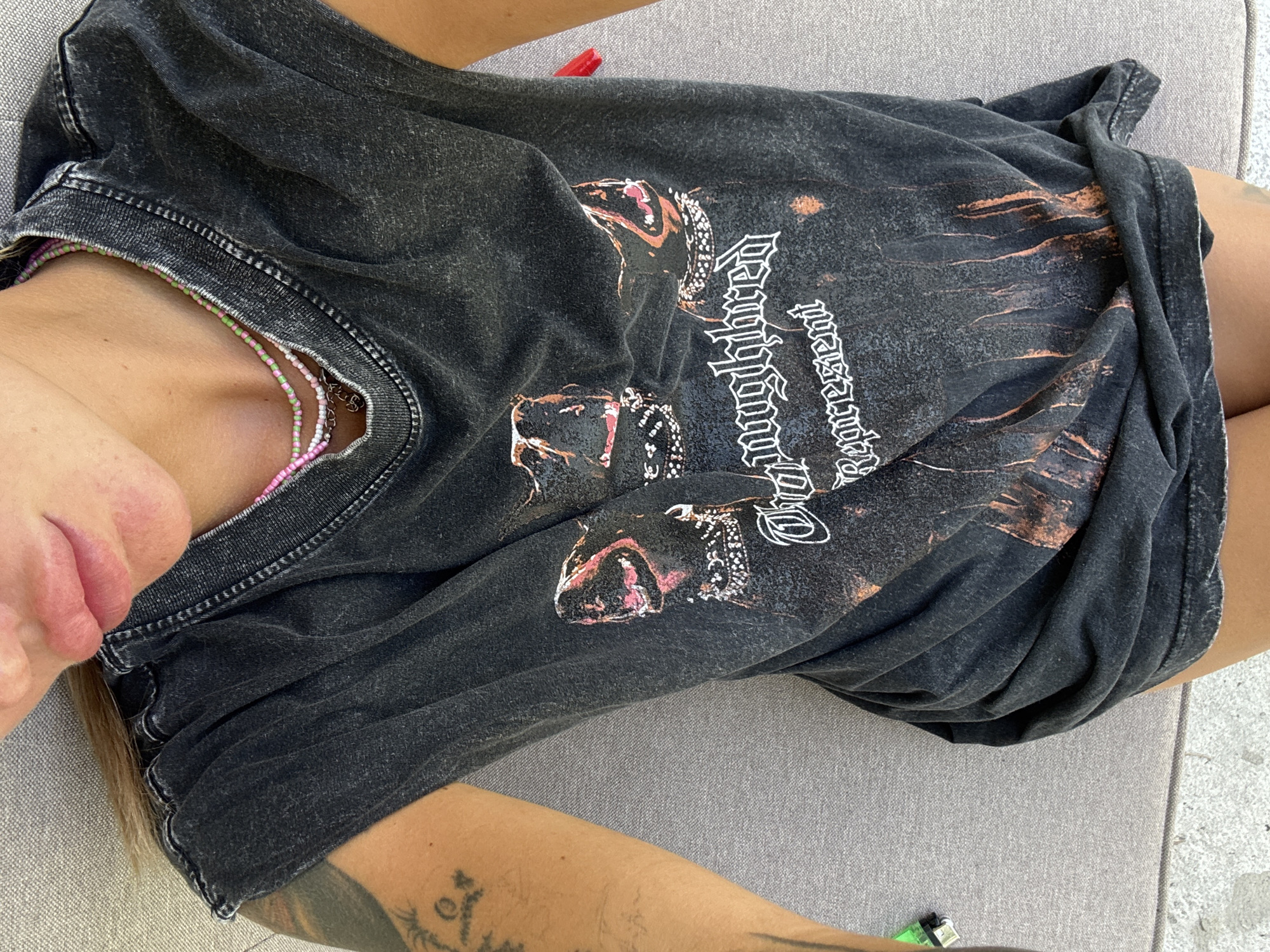 The image depicts a person lying on a gray surface. They are wearing a black t-shirt with a graphic print and beaded necklaces. The person has tattoos on their arm. The environment is not distinctly identifiable. The location of Choeng Thale, Thailand is not evident from the image.