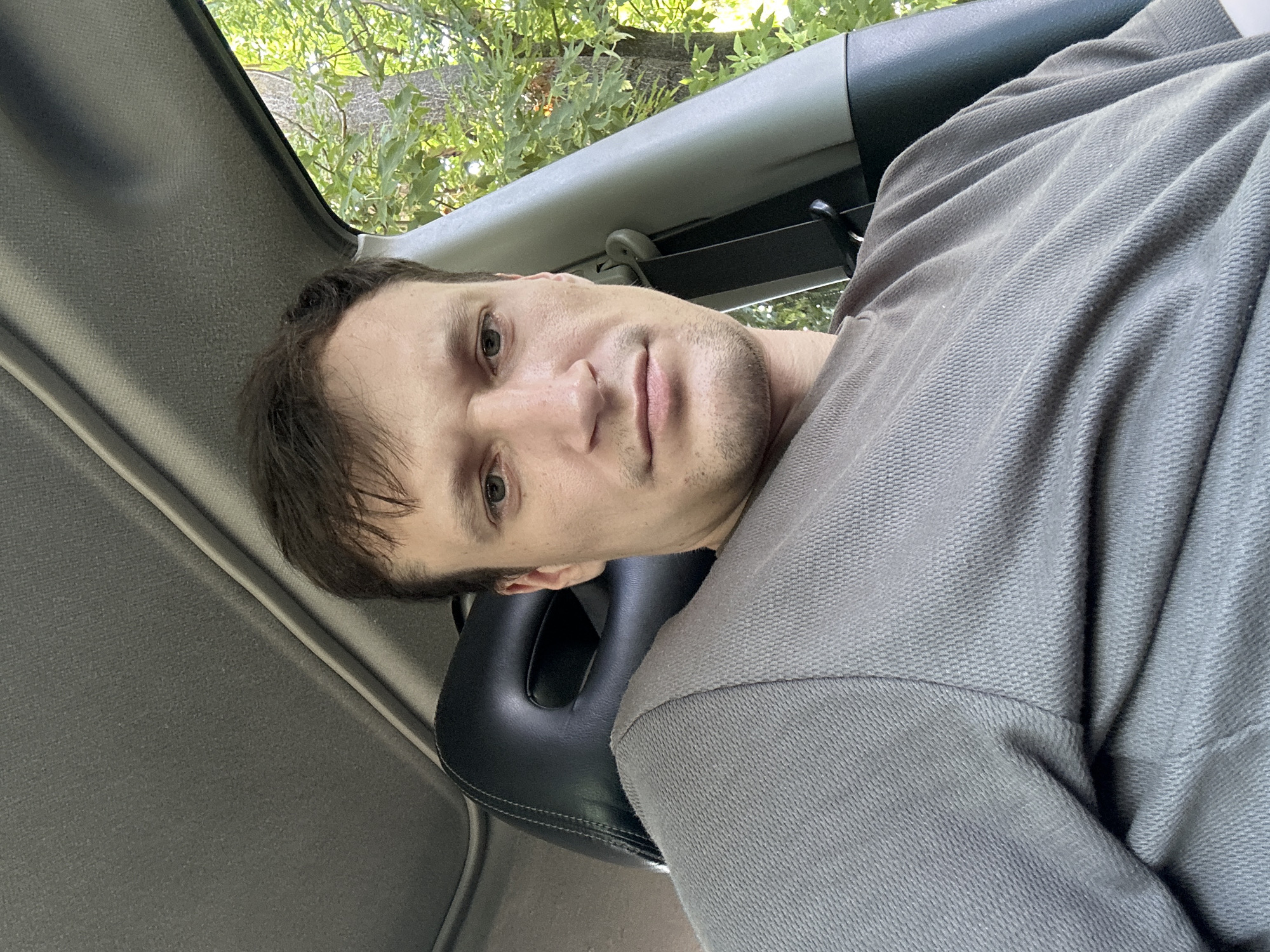 The image is a selfie taken inside a car, with the passenger seat headrest and a portion of the car's interior visible. The subject is a man with short brown hair and light skin, wearing a grey t-shirt. He is looking directly at the camera with a neutral expression. Outside the car, through the window, a tree with green leaves is visible, suggesting an outdoor, possibly rural or suburban, setting. The lighting indicates it is daytime. No specific location in Makiv, Ukraine, can be determined from the image.