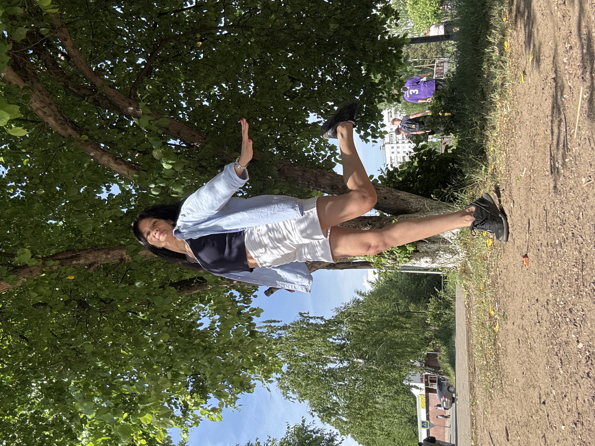 A young woman is standing in a park. The woman is light-skinned and has long black hair. She is wearing a dark cropped shirt, a light-blue button-down shirt, and white shorts. Her arms are extended out in front of her. The woman is wearing sneakers. The park is green and lush, with tall trees overhead. The sky is blue, with wispy clouds. A walking path is visible. In the distance, people are wearing purple jerseys. A building can be seen in the distance. The image is taken outdoors, and the lighting is bright.
