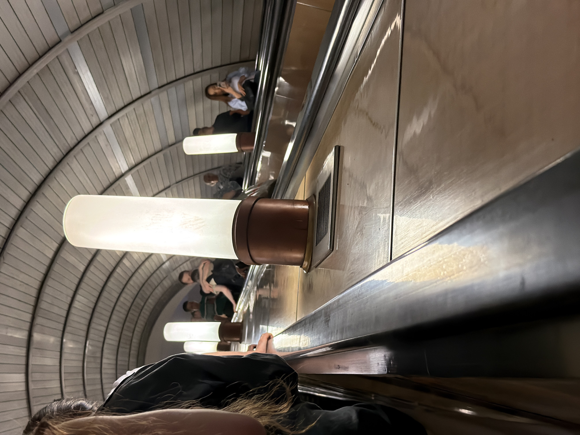 The image depicts an escalator in the Moscow Metro system, specifically at the Komsomolskaya station, known for its ornate architecture. The escalator's ceiling is a ribbed, arched structure made of metal. Along the side of the escalator, there are tall, cylindrical white lights with copper-colored bases. Several people are visible on the escalator, moving upwards. In the foreground, on the left, a person's shoulder and part of their hair are visible. Further up the escalator, a woman in a light blue shirt is holding a phone. Other individuals are present but less distinct. The escalator steps are made of metal with a reflective surface, and the side panels are also metallic, reflecting the surrounding lights and structures.