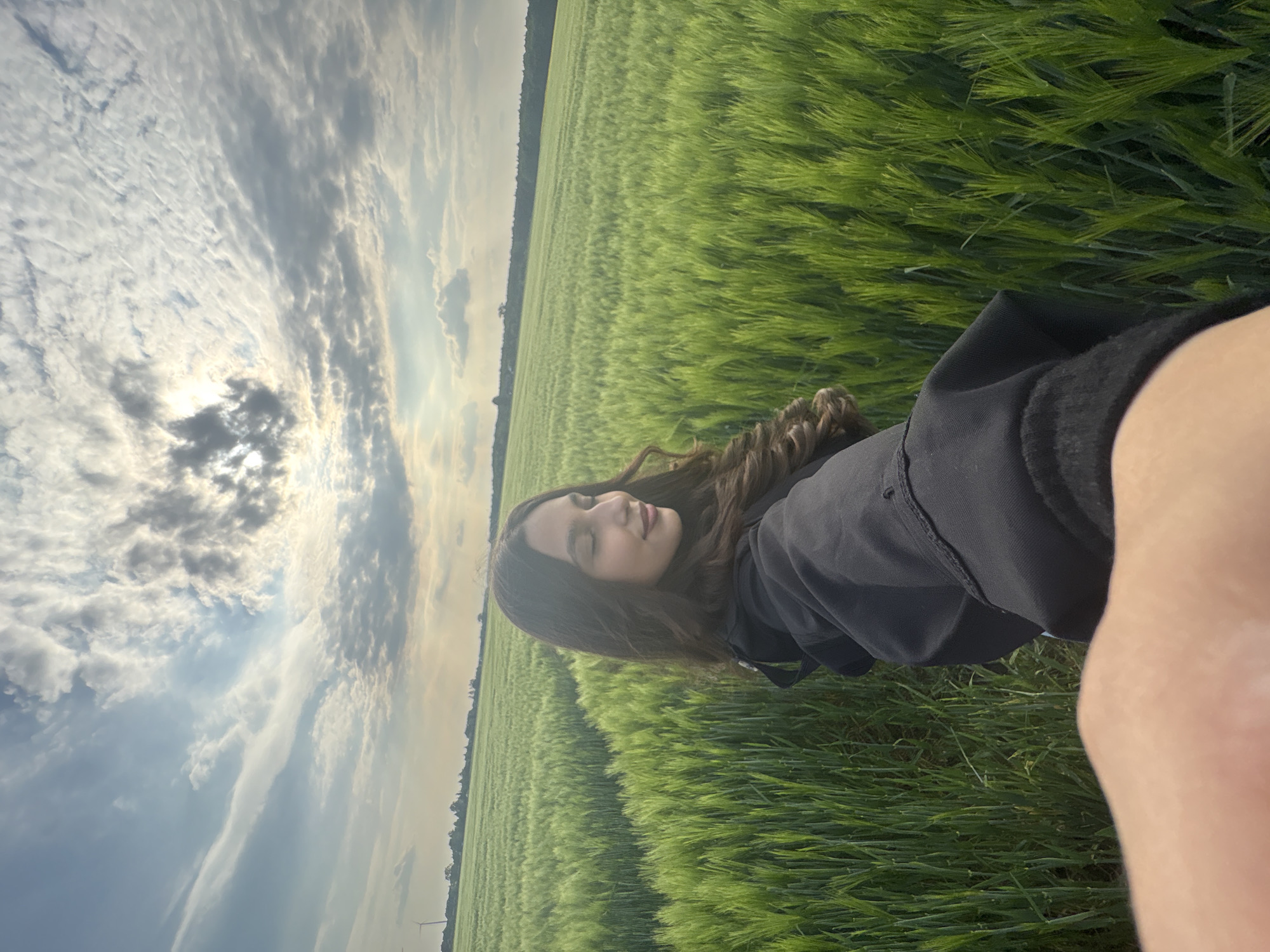 A selfie shows a girl lying in a green field under a cloudy sky with the sun peeking through. She has long, curly brown hair and is wearing a dark jacket or shirt. The field appears to stretch out extensively. The angle and lighting suggest the photo was taken around sunset. The location is Wegberg, Germany.