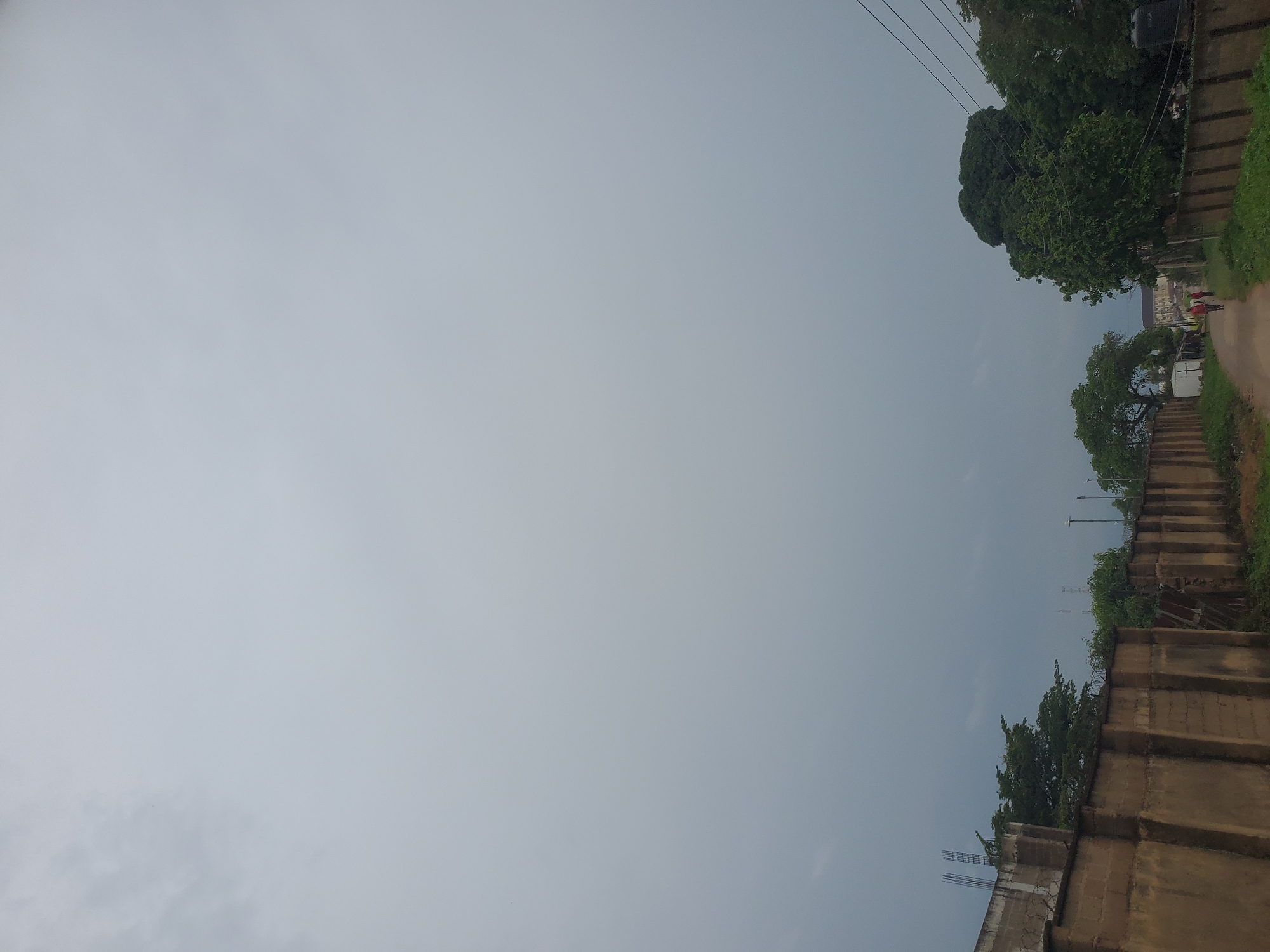 The image depicts a cloudy sky over a dusty, unpaved road in Jos, Nigeria. On the left side of the road, a low concrete wall runs along the edge of a property. Behind the wall, several structures are visible, including one with exposed rebar, suggesting ongoing construction. To the right of the road, a dense collection of trees forms a natural barrier, with a few scattered utility poles and electrical wires running overhead. Further down the road, two individuals are walking away from the camera. The overall atmosphere is subdued, likely due to the overcast weather.