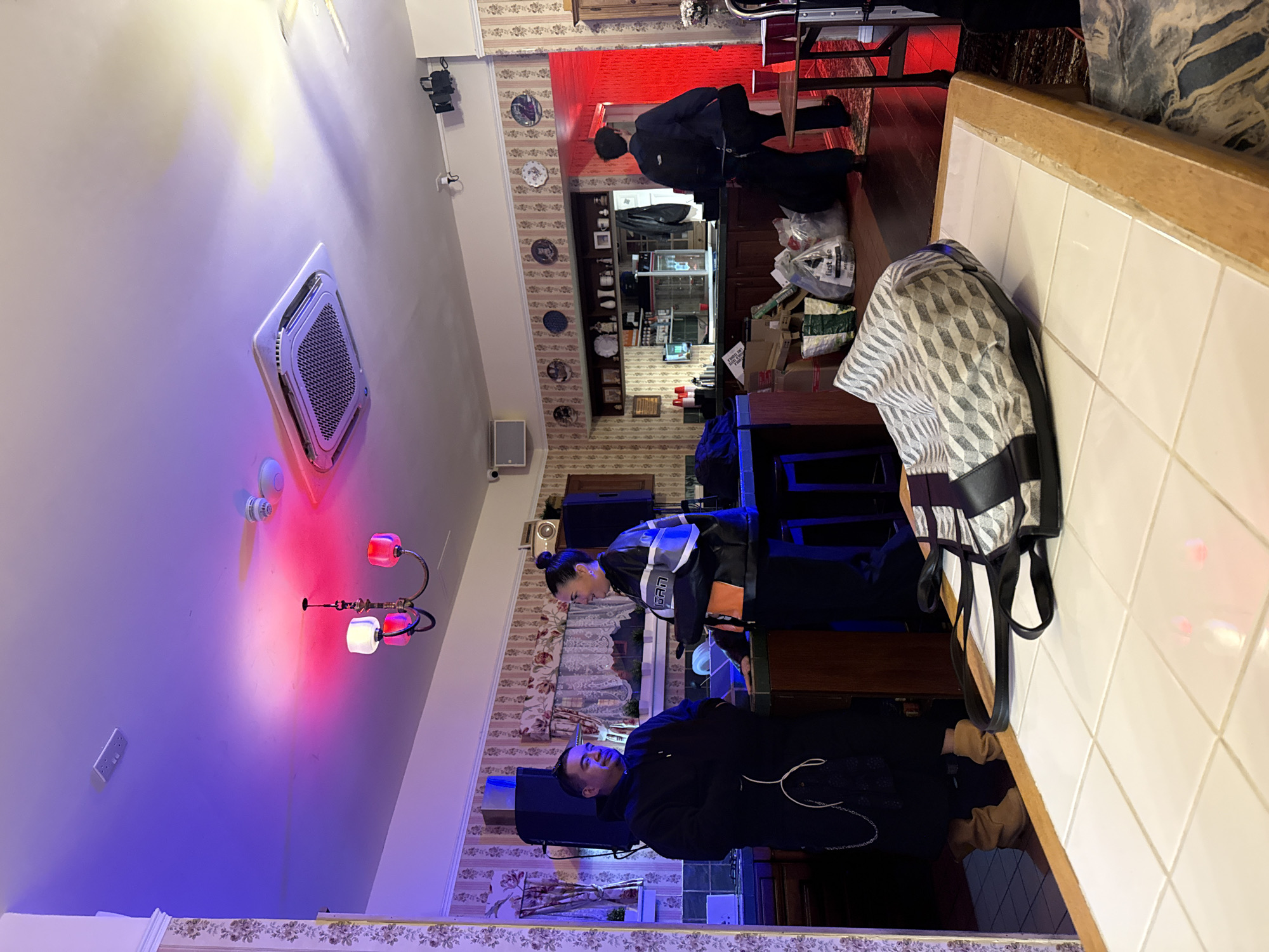 The image captures an indoor scene within a commercial establishment, likely a bar, cafe, or restaurant in London, United Kingdom. The room features a mix of traditional and modern elements, including patterned wallpaper, decorative plates mounted on the walls, and wooden counters.
Three individuals are present. In the foreground, a man wearing a dark jacket or hoodie stands looking towards the camera. Behind him, a woman with dark hair tied back is standing, wearing a dark jacket with white and light blue patterns, and looking downwards. Further back, behind a service counter, a third person, a man, is bending over, appearing to be at work.
Key objects include a white-tiled counter with a wooden edge in the foreground, from which a geometric-patterned (black, white, and grey) bag is hanging. On the opposite side of the room, there is a bar or counter area with shelves displaying various items. Near this counter, several bags and cardboard boxes are visible, with one clear plastic bag bearing the partially discernible text "first lie" or similar. Overhead, an air conditioning unit and a decorative light fixture with red and white/pink shades contribute to the lighting.
The setting is primarily illuminated by artificial light, suggesting evening or night. White overhead lights combine with prominent red and blue/purple accent lighting, casting distinct colored glows on the walls and ceiling. The overall ambiance is casual.