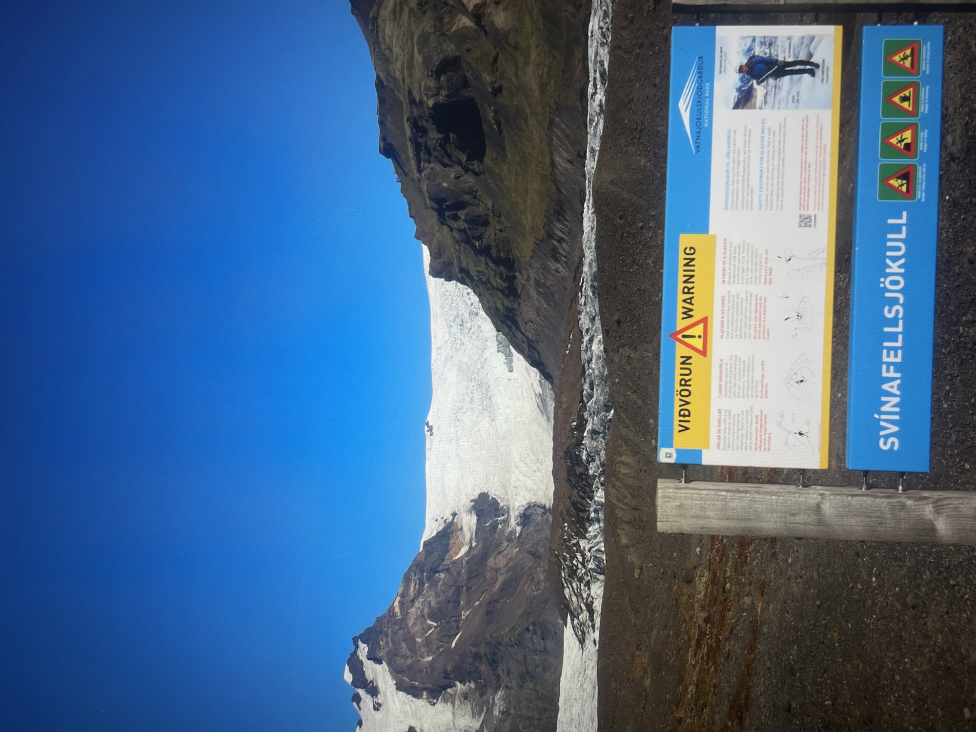 Here is a description of the image:
The image shows a glacier in Iceland, under a clear blue sky. To the right is a warning sign about the dangers of glaciers and ice caves, titled "SVÍNAFELLSJÖKULL". The sign includes safety information for glacier walks and cautions about hazards such as crevasses. The terrain near the sign appears to be rocky and gravelly. A portion of the mountains is visible in the background.