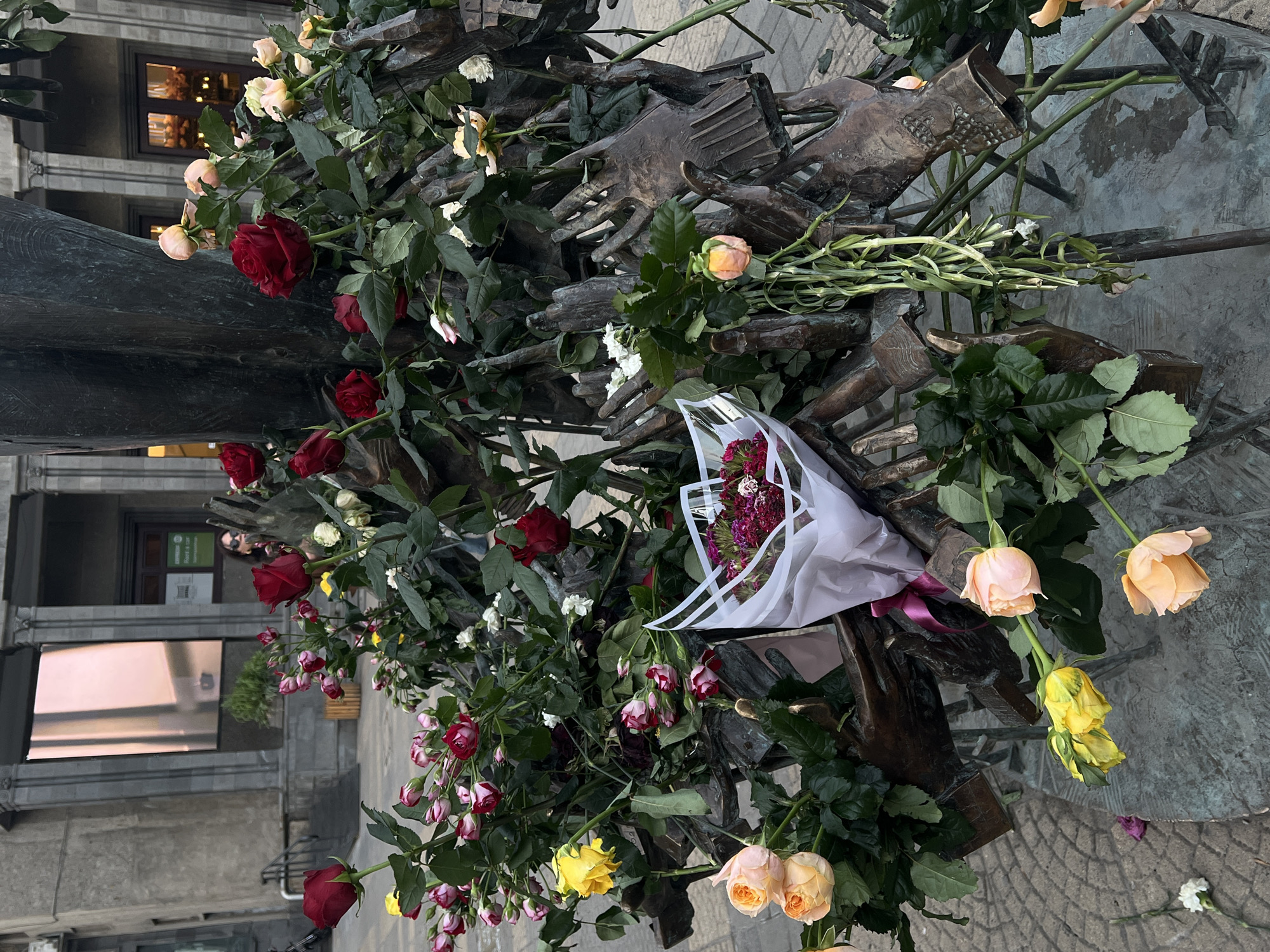 A bronze sculpture of interlocking hands is covered in roses of various colors, including red, yellow, white, and pink. A bouquet wrapped in white paper is nestled among the hands. The sculpture sits in a paved area with buildings in the background. The location is the city of Unknown, Unknown.