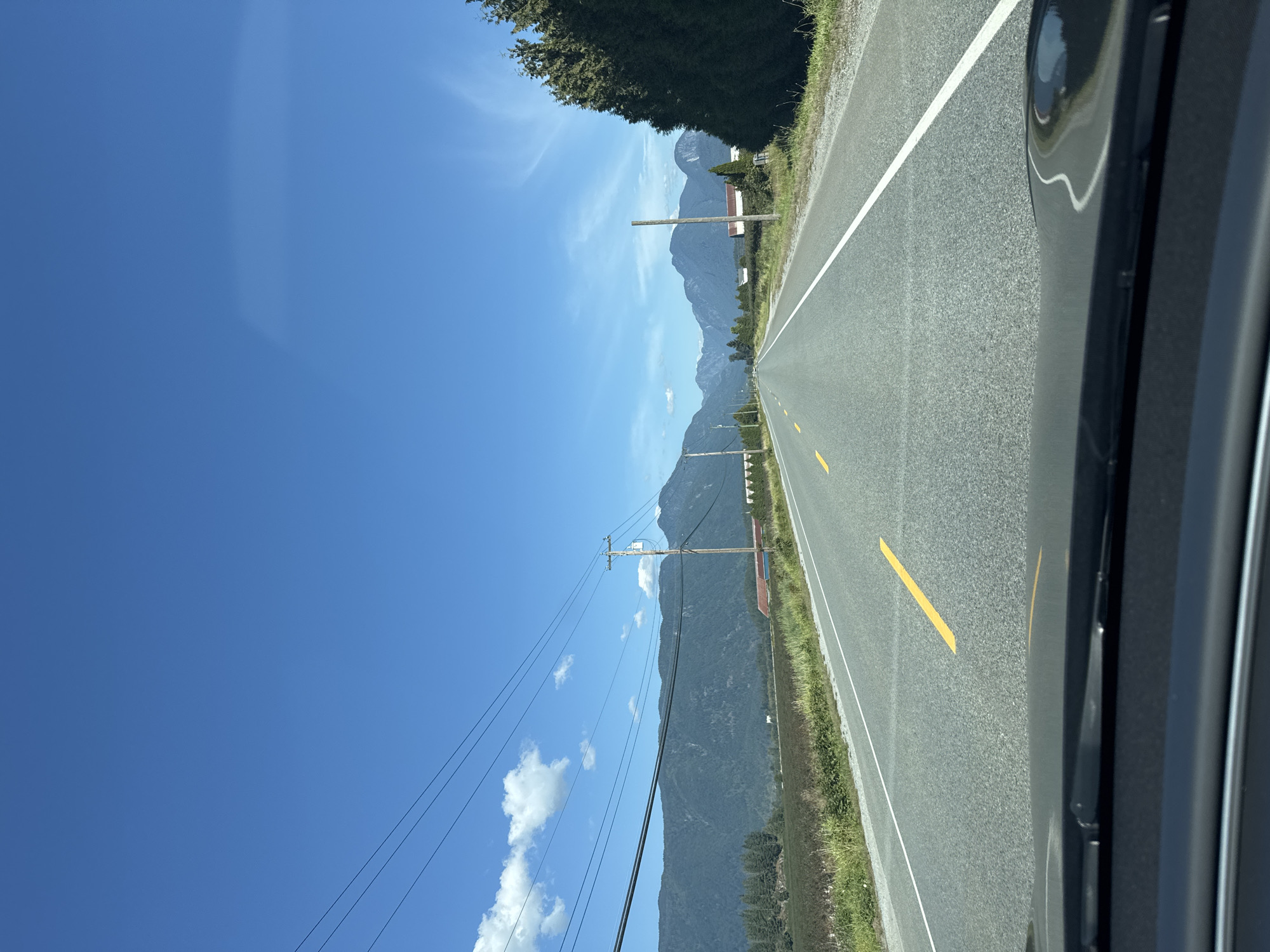 The image shows a view from a vehicle driving on a two-lane road in Pitt Meadows, Canada. The road is asphalt with yellow and white lane markings. On the left side of the road, there is a field of crops, followed by a line of trees and then a range of mountains. Power lines run parallel to the road on the left side. On the right side of the road, there are trees and some buildings. The sky is clear and blue with a few scattered clouds. The sun appears to be shining, casting shadows on the road and landscape. The scene appears to be rural and mountainous, with signs of agriculture and infrastructure.