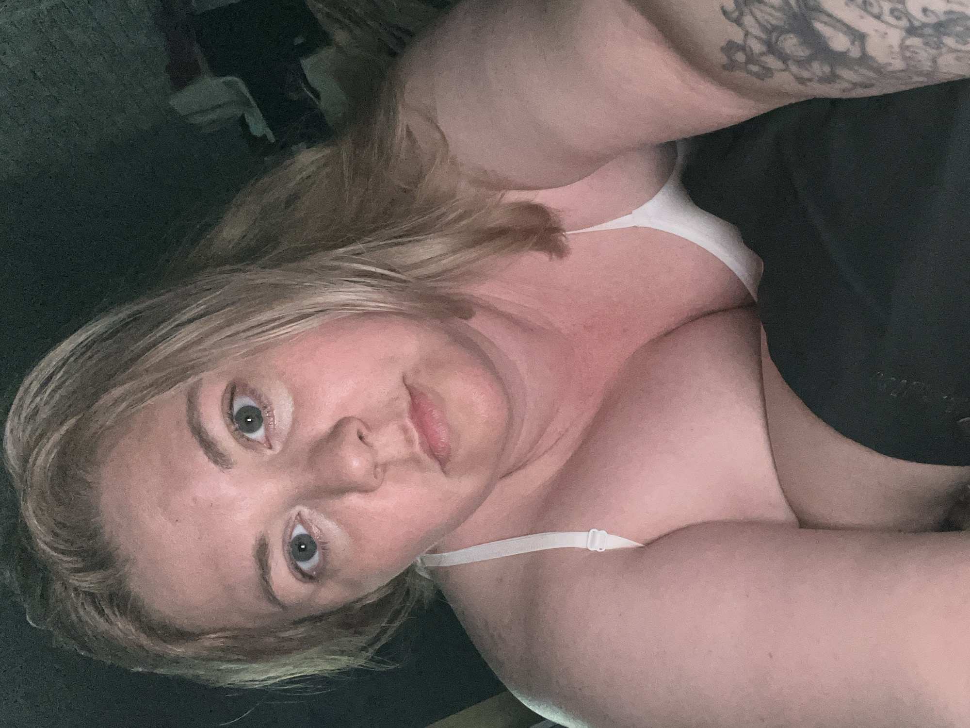 A fair-skinned individual, likely female, with blonde hair and light-colored eyes, is depicted in a close-up, vertical shot. The person is lying down, with their head positioned towards the lower left of the frame and their body extending upwards to the right. They are looking directly at the camera with a neutral to slightly pensive expression.
The individual is wearing a white bra or camisole top with thin straps. A significant portion of their upper body is visible, including their décolletage, shoulders, and arms. A prominent, dark-colored tattoo with intricate designs is visible on the upper right arm or shoulder.
The background is dark and indistinct, suggesting an indoor setting with low lighting, possibly a bedroom or living area. Some shadowy, undefined shapes can be seen in the upper left corner of the background, hinting at furniture or other objects. The lighting is soft and appears to originate from the front, illuminating the person's face and upper body while casting gentle shadows. The overall ambiance is intimate and personal. There is no visible text in the image. The time of day is not explicitly clear but the dim lighting might suggest evening or a room with closed curtains. The location is an indoor, private space, with no specific cues relating to Vesyoloye, Russia.