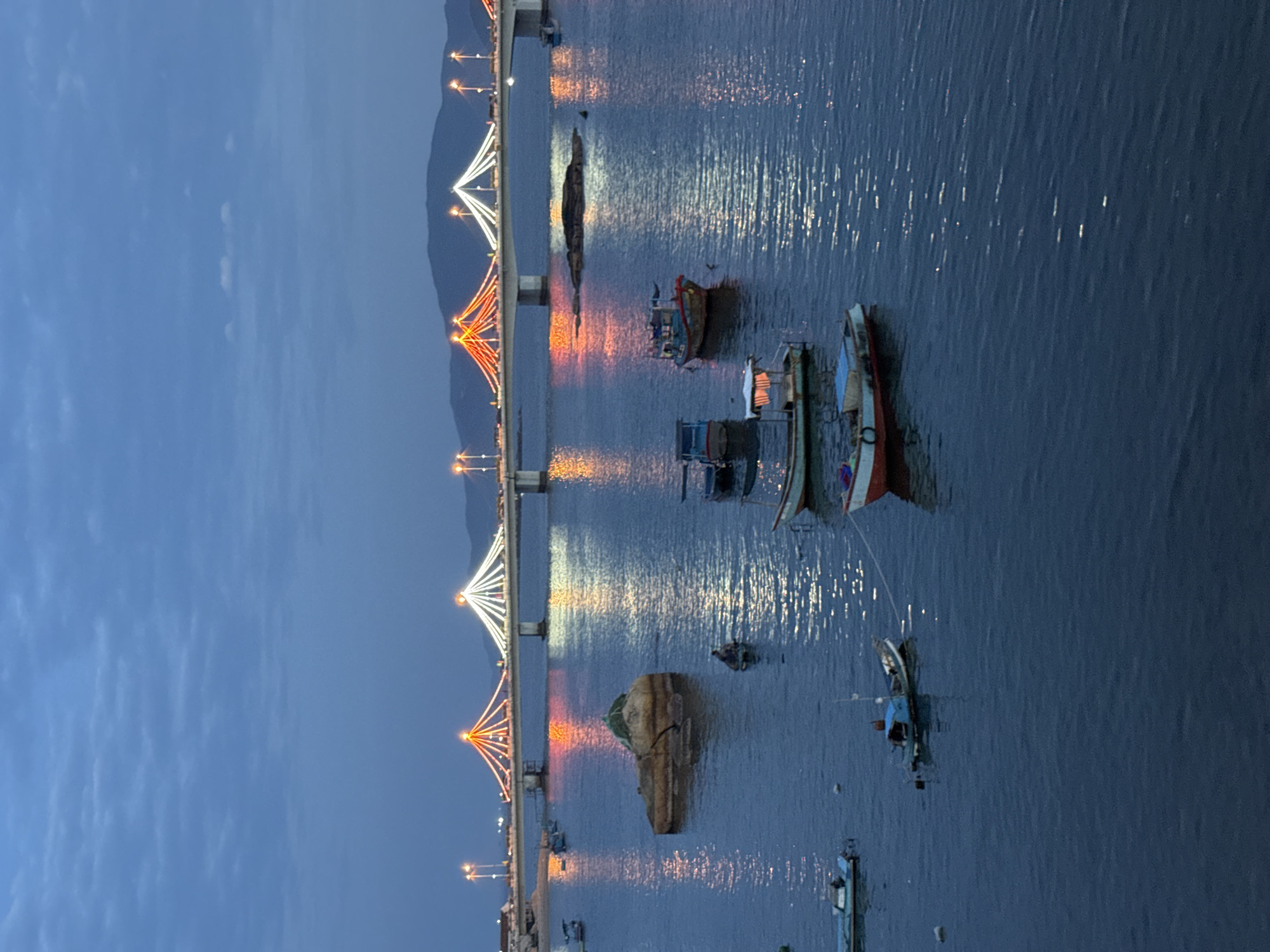 The image depicts a twilight scene of a bridge illuminated with decorative lights, spanning a body of water in Nha Trang, Vietnam. The bridge, likely the Tran Phu Bridge, is adorned with strings of white and orange lights that create a festive atmosphere. Reflections of these lights shimmer on the calm water's surface.
Several boats are anchored in the water. In the foreground, a few small fishing boats are visible, some appearing weathered. One boat is a light blue and white vessel with a covered cabin, while another is a larger, more traditional fishing boat with a colorful hull. In the middle ground, a larger, tan-colored rock formation rises from the water, with a single person seated on it. Further out, more boats are scattered across the water.
The sky above is a deep blue, with wispy clouds suggesting the transition from day to night. Hills are visible in the distance behind the bridge. The overall impression is one of a tranquil evening in a coastal city, with the bridge serving as a prominent landmark.