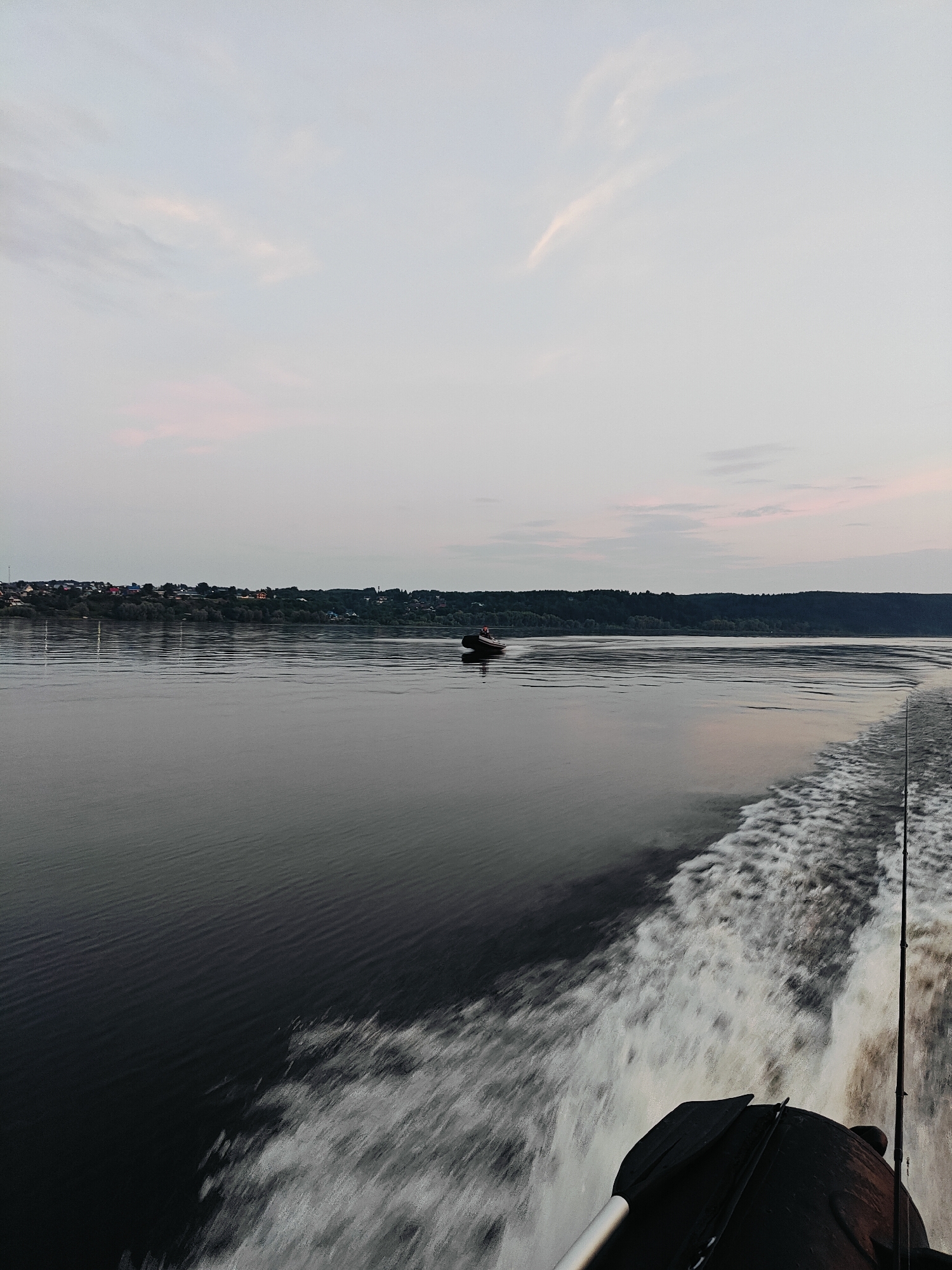 The image depicts a wide, calm body of water, likely a river, at dusk. A small, dark boat with a person wearing a red hat is visible in the distance, moving across the water. The wake of a larger boat, from which this photo is taken, churns white water on the right side of the frame, indicating forward motion. In the background, a tree-lined shore with a cluster of buildings suggests a settlement, possibly the city of Noviy, Russia. The sky is a muted gradient of pale blue and pink, with wispy clouds.