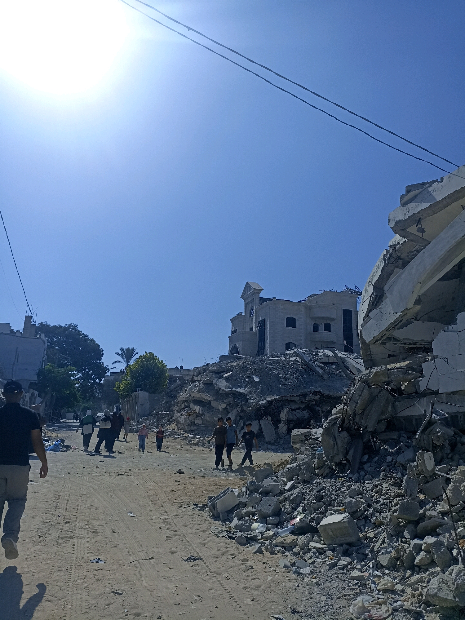 A bright, sunny day in Gaza City, Palestinian Territories, reveals a scene of widespread destruction. The foreground is a dirt road with tire tracks, strewn with rubble and debris. To the right, a large, collapsed building dominates the frame, its concrete structure broken and scattered. In the mid-ground, a group of people, including children, walk along the road amidst the ruins. Behind them, another building, though damaged, stands partially intact. Power lines crisscross the clear blue sky, with the sun shining intensely, casting long shadows. The overall impression is one of devastation and the resilience of daily life continuing in its aftermath.