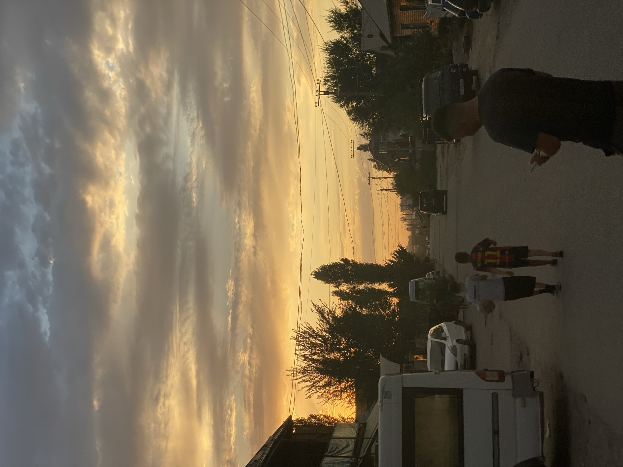 The image depicts a residential street in Bishkek City, Kyrgyzstan, during a sunset. The sky is filled with dramatic clouds painted in hues of orange, yellow, and grey. Several children are visible on the street; two young boys in the foreground are walking away from the camera, one carrying a basketball and the other wearing a soccer jersey. A man in the foreground is also walking away, looking at his phone. Various vehicles, including a white van, a white car, and a dark SUV, are parked or driving on the street. Buildings line the sides of the road, and trees create silhouettes against the vibrant sky. Power lines and utility poles are prominent features of the urban landscape. The overall scene suggests a typical evening in a residential area, with people and vehicles present as dusk approaches.