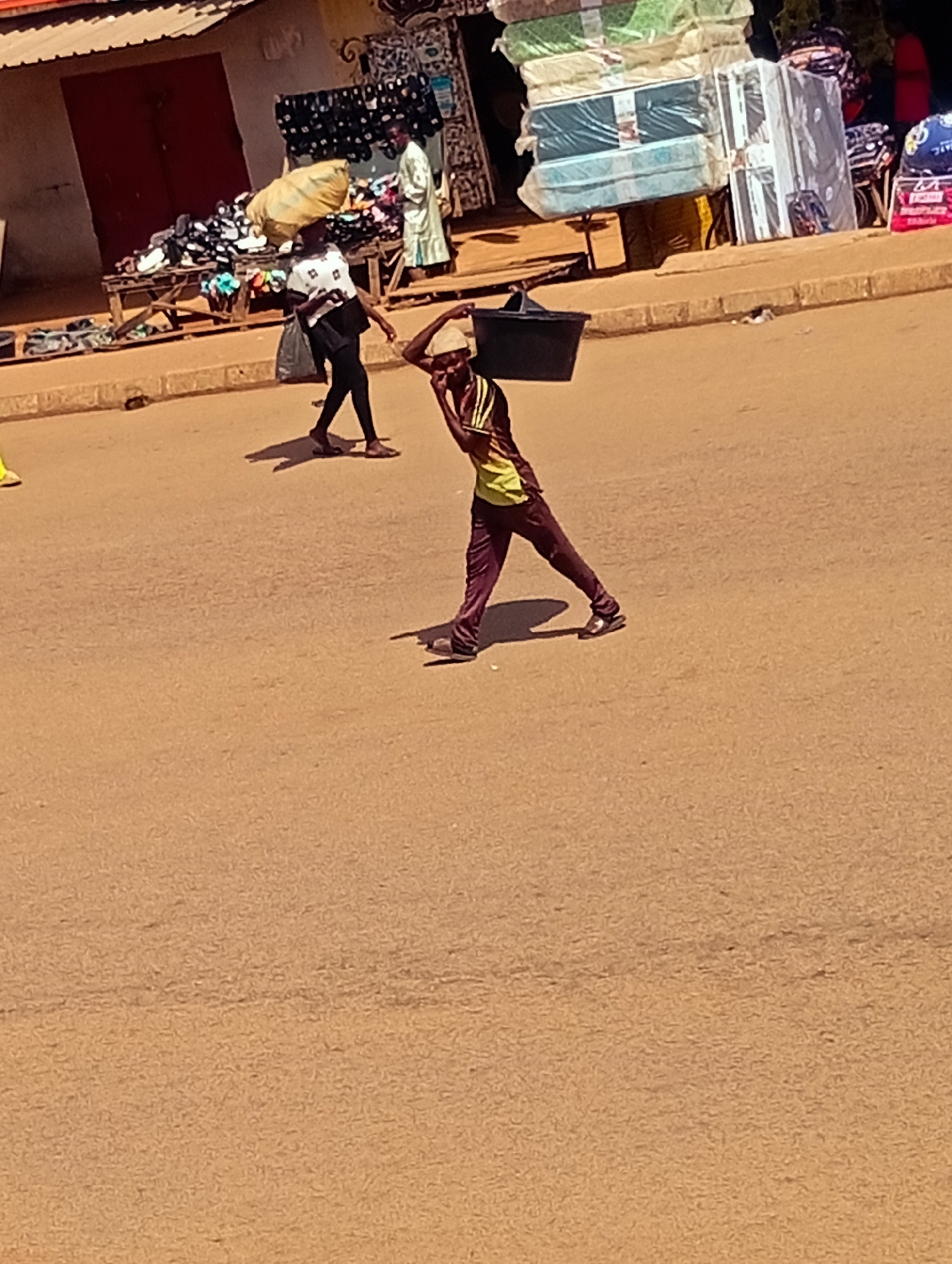 In Bukuru, Nigeria, a person walks along a street carrying a black bucket on their head. The person is wearing a yellow and green striped shirt and maroon pants. The street is paved and appears to be a commercial area with shops lining the background. One shop has mattresses stacked outside, wrapped in plastic. Another shop displays shoes on a wooden stand. Several other people are visible in the background, including a woman carrying a large bag on her head. The scene suggests a busy marketplace during the daytime, with people engaged in commerce and transport. The lighting indicates it is daytime, and the warm tones suggest a sunny climate.
