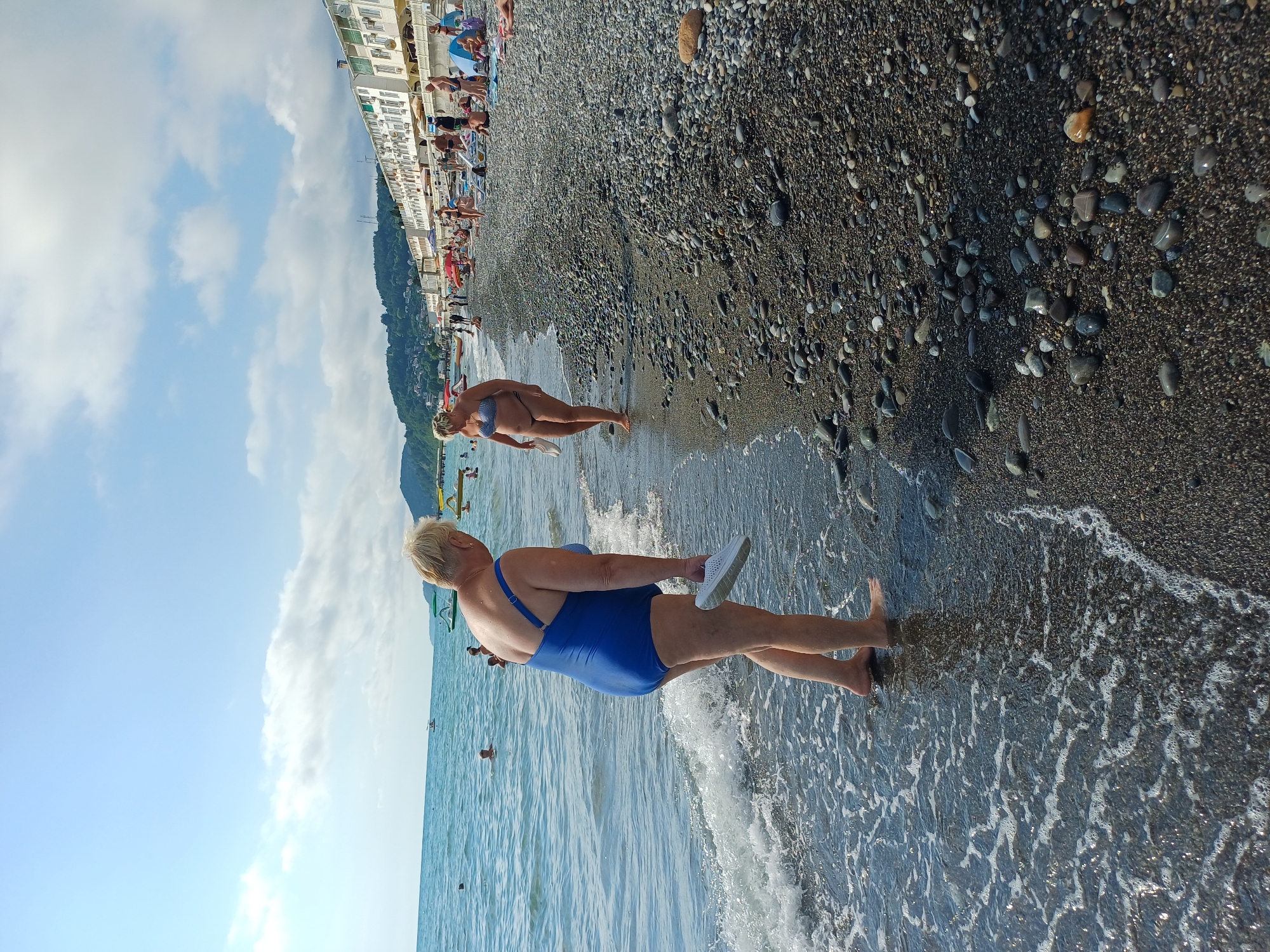The image depicts a beach scene in Sochi, Russia. In the foreground, an older woman wearing a blue one-piece swimsuit walks towards the water's edge, holding a white sandal in her right hand. Her feet are partially submerged in the shallow waves. Further down the beach, another woman in a bikini walks along the shoreline. The beach is covered in dark pebbles. In the background, several people are sunbathing on lounge chairs, some under umbrellas. Buildings with white facades and balconies are visible along the coastline, with a green, tree-covered hillside rising behind them. The sky is blue with scattered white clouds, and the sea is calm. There are also some boats visible in the water. The scene appears to be a typical summer day at a popular beach resort.