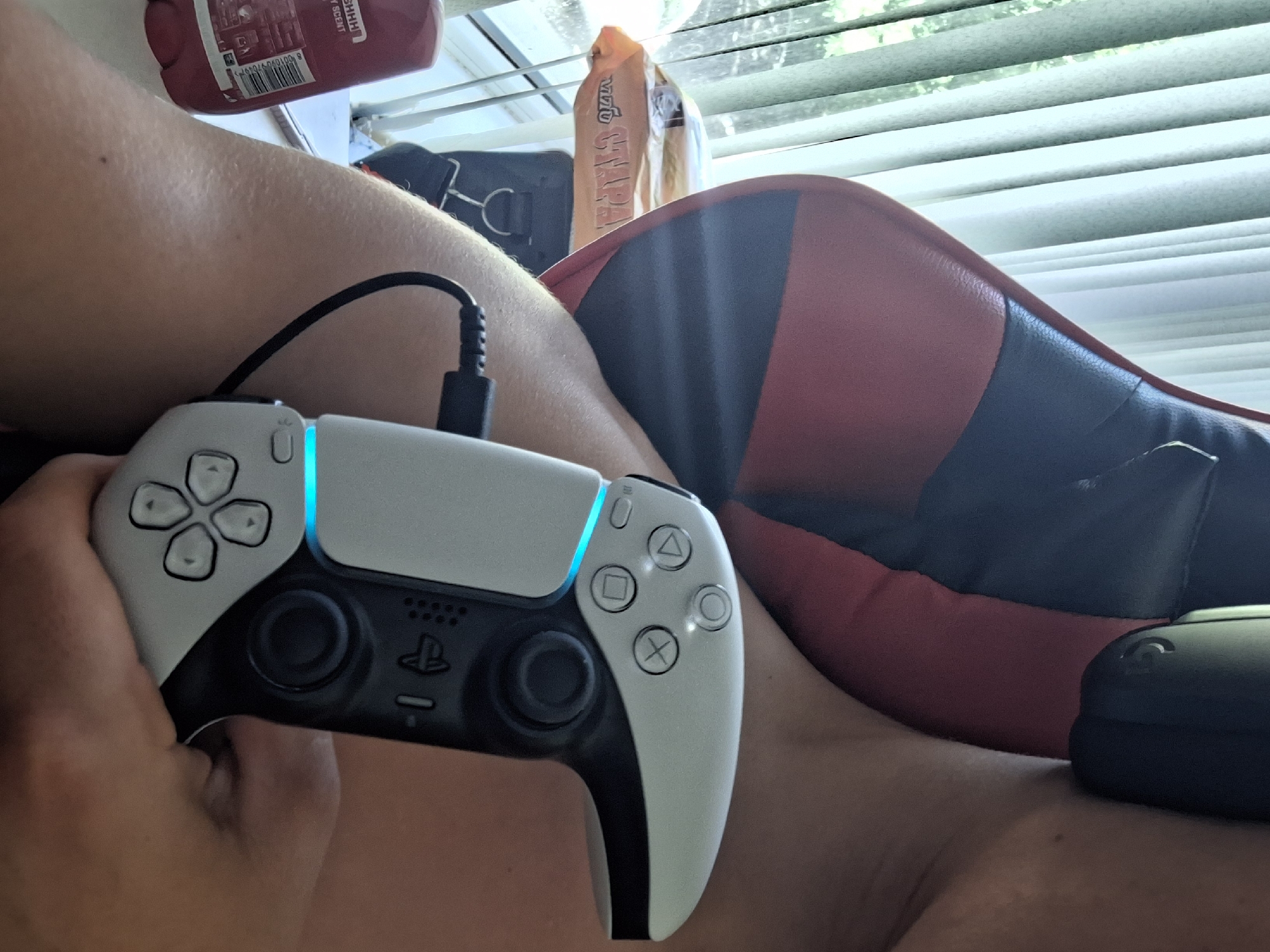 The image shows a close-up of a person holding a white PlayStation 5 controller, which is plugged in via a black cable. The controller has illuminated blue accents. The person's arm and shoulder are visible, and they appear to be leaning back against a red and black gaming chair. In the background, there are vertical blinds allowing light to enter, and a roll of what appears to be toilet paper is visible on a surface. There is also a red bottle, possibly of body wash, with a label that includes the word "SCENT" and a barcode. The scene is indoors, and while there are hints of greenery visible through the blinds, the specific location within Varna, Bulgaria, cannot be determined from the provided image. The overall impression is a casual setting, likely a home environment, where the person is preparing to or is currently gaming.