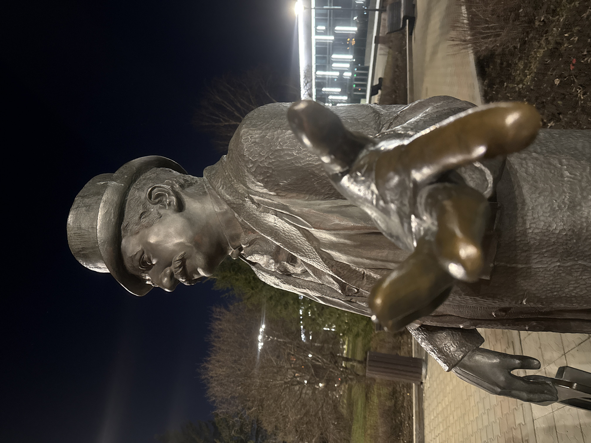 A bronze statue of a man wearing a hat and suit is the focal point of the image, taken at night in what appears to be Yekaterinburg, Russia. The man is depicted with his hand outstretched, as if offering a handshake or inviting someone closer. The statue is situated on a paved area, and the background features trees, shrubs, and illuminated buildings. The scene is lit by artificial lights, highlighting the statue and its surroundings.