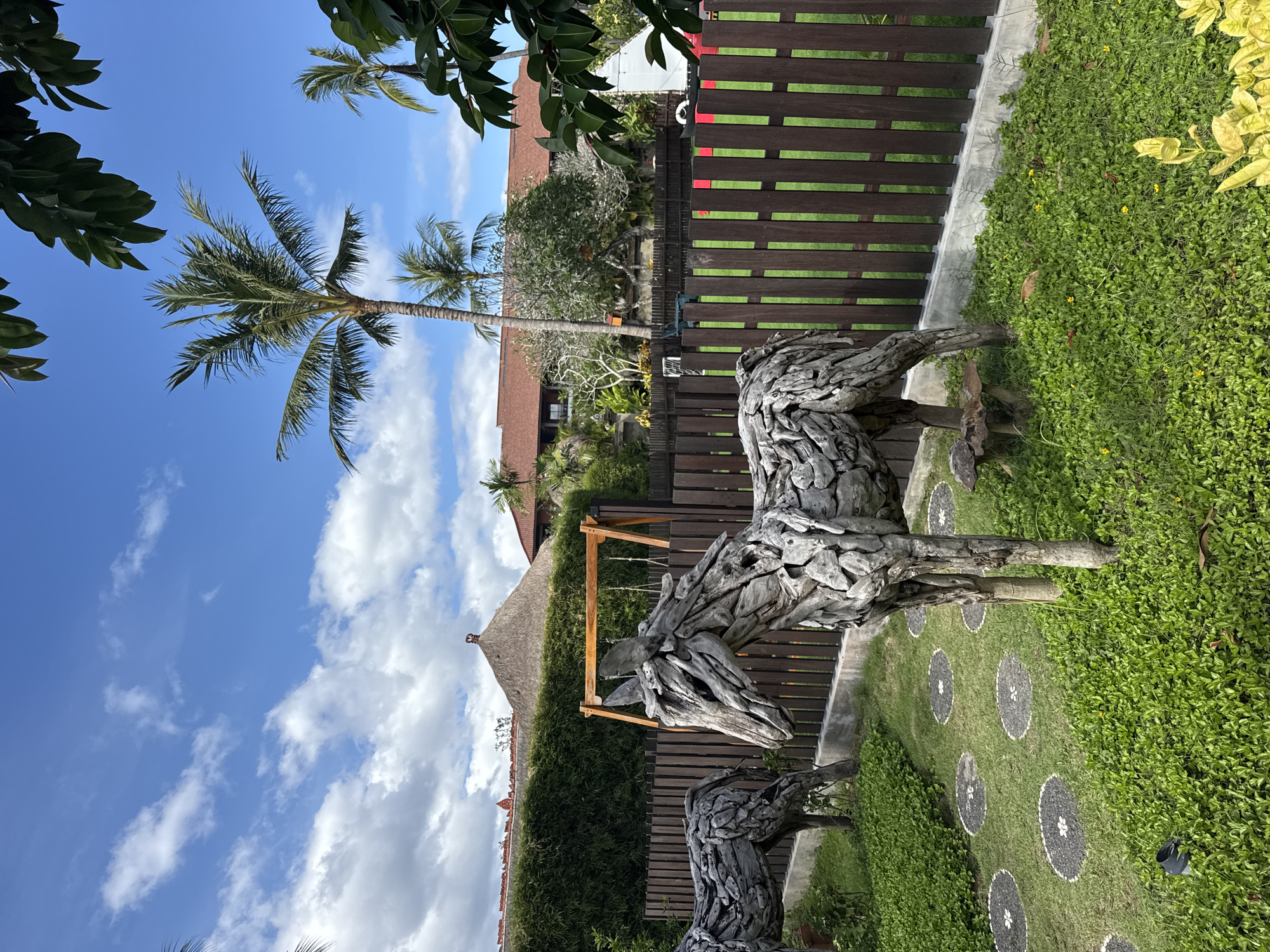 The image depicts an outdoor scene in Kuta Selatan, Indonesia. The foreground features two wooden sculptures of horses, constructed from driftwood. They stand on a lawn adorned with a pathway consisting of circular stone patterns. Behind the sculptures, a wooden fence runs along the property line. A triangular roof structure with a thatched top is visible beyond the fence, partially obscured by greenery.
In the background, a tall palm tree extends into a blue sky with scattered white clouds. Further back, rooftops of buildings are visible amidst the lush vegetation. The scene is bathed in daylight.