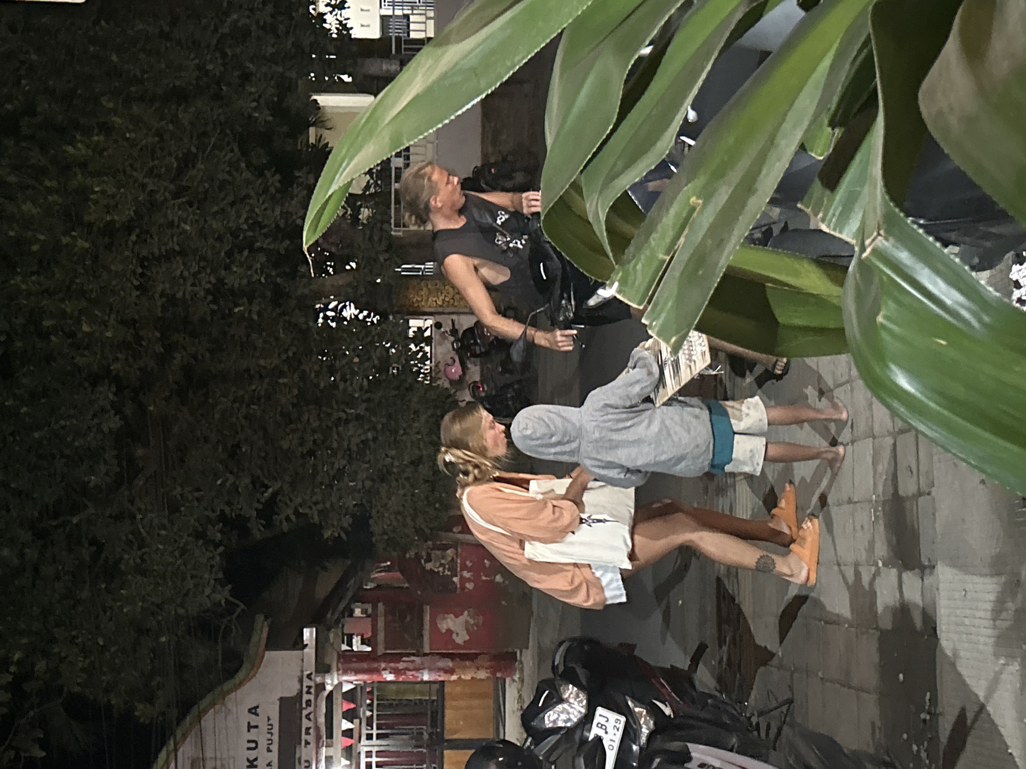 The image depicts a street scene at night in Kuta, Indonesia. In the foreground, a large, lush green plant partially obscures the view. To the left, a motorcycle is parked, showing its front tire, license plate "BJ 01-29", and handlebars. Further back, a young child wearing a grey hoodie and shorts stands on a tiled sidewalk, looking down at something in front of them. Beside the child, a woman with blonde hair tied back, wearing an orange cardigan over a white skirt and orange sandals, is bent over, interacting with the child and holding a white tote bag. She has a tattoo on her left calf. To the right, a man with blonde hair, wearing a black sleeveless shirt and dark pants, is seated on a motorcycle, leaning forward as if preparing to ride. The background shows a building with signage that includes "KUTA" and "LA PUJUT", suggesting a location in Kuta, Bali, Indonesia. Trees with dark foliage are visible overhead, and other motorcycles are parked in the distance. The ground is wet, with reflections indicating recent rain.
