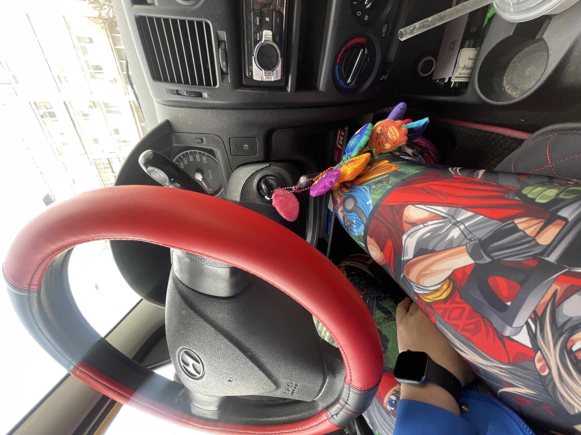 The image captures the interior of a Hyundai vehicle, consistent with a first-generation Hyundai Getz, from the driver's perspective, located in the city of Chaikovsky, Russia.
In the foreground, the lower half of a person's body is visible, seated in the driver's seat. The individual is wearing leggings or trousers extensively patterned with vibrant, multi-character anime or manga artwork. A left forearm, clad in a blue long-sleeved garment, rests on the left thigh, displaying a black smartwatch with a square face on the wrist.
The black steering wheel, featuring the Hyundai logo, is partially covered by a red and black faux leather grip. Beyond the steering wheel, the dark dashboard includes an instrument cluster with an analog speedometer calibrated up to 220 km/h and a 'TRIP' button. The vehicle's ignition key is inserted, adorned with an elaborate cluster of colorful keychains, including a prominent pink fluffy pom-pom, several smaller multi-colored fluffy pieces, and a distinct orange charm depicting a smiling monster.
To the right, the center console houses a car radio unit (partially branded "MIC"), air conditioning vents, and rotary climate control dials. Below these, a cup holder contains a clear plastic cup with a straw. An adjacent storage compartment holds a small, dark-colored bottle and a white rectangular object, possibly a packet of cigarettes. The floor of the vehicle is dark and exhibits some minor debris.
Through the windshield, a light-colored, multi-story building, characteristic of urban residential or commercial architecture, is visible under an overcast sky. The building facade features numerous windows and several externally mounted air conditioning units. The precise street address within Chaikovsky, Russia, cannot be determined from the visual information.