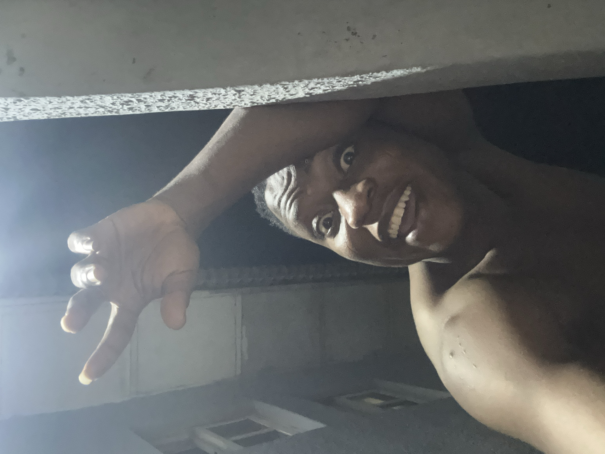 A dark-skinned individual, appearing male, is captured in a close-up, low-angle shot, seemingly a selfie, within a dimly lit urban or residential environment consistent with an area in Ikorodu, Nigeria. The individual is shirtless, exposing the right shoulder, upper arm, and a portion of the chest. Small, raised skin irregularities are visible on the anterior aspect of the right shoulder. The facial expression is characterized by wide-open eyes, raised eyebrows, and an open mouth revealing a full set of white teeth, conveying an exaggerated or playful grimace. The individual's right arm is extended upwards, with the hand displaying a gesture where the index and pinky fingers are extended, and the middle and ring fingers are curled, with the thumb tucked. The distal ends of the extended fingernails appear lighter in color. The left arm is bent, with the forearm resting against a light-colored, textured concrete pillar or wall that occupies the right side of the frame. The background on the left reveals a portion of a building structure with a rectangular window-like opening. A prominent light source originating from the upper left illuminates the subject's face and raised hand.