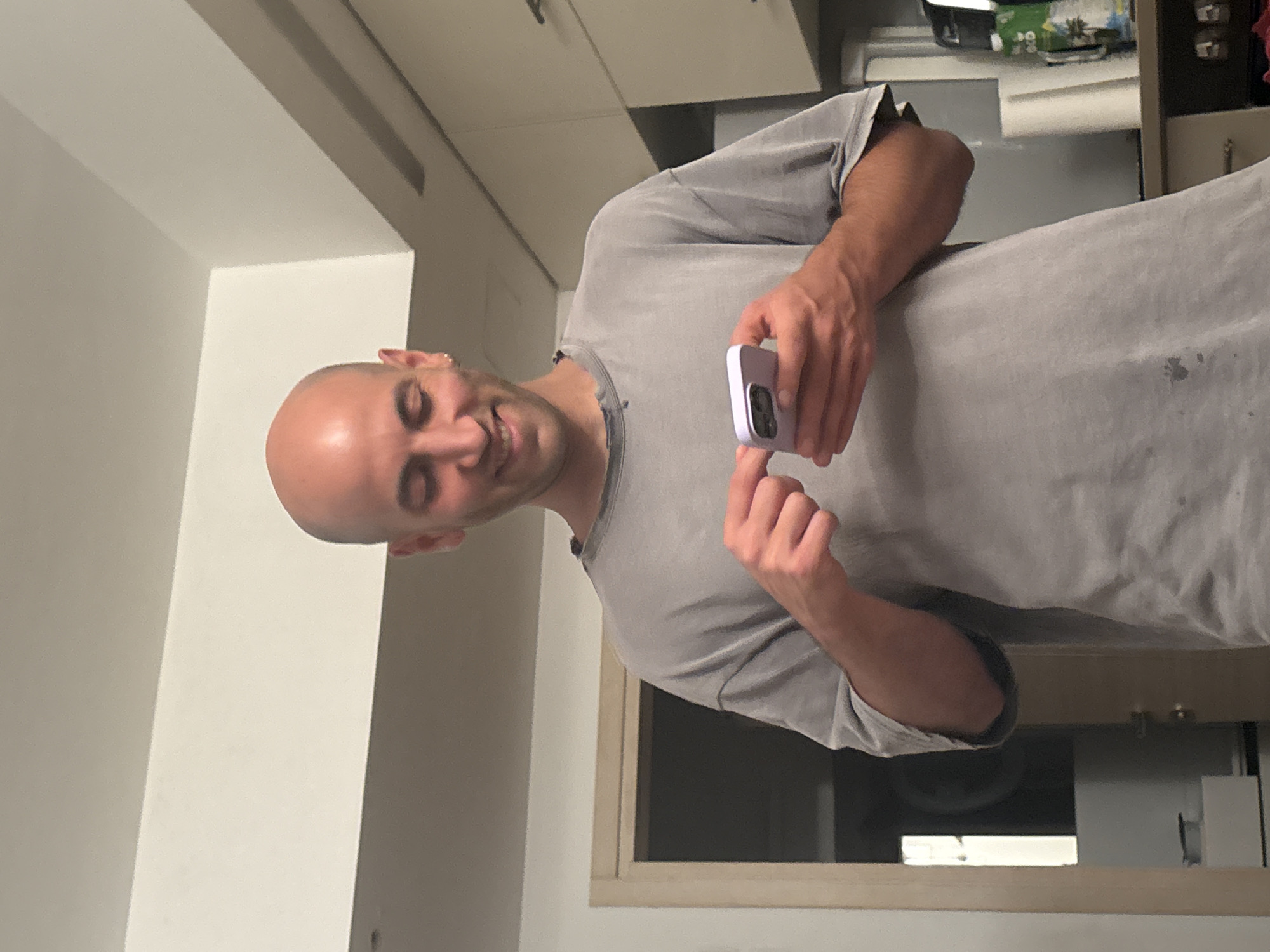 A male individual is depicted indoors in a residential setting in Tel-Aviv, Israel, likely within a kitchen or an area adjacent to one. The man, who is bald and wears a small gold-colored hoop earring in his visible left ear, is attired in a light grey, possibly distressed or oversized, short-sleeved t-shirt with several dark stains on the lower front. He is observed smiling while looking down and actively interacting with a smartphone. The phone, encased in a light purple protective cover, is held in his left hand, while his right index finger is positioned on its screen. The background to the right reveals light-colored kitchen cabinetry with dark handles, a roll of white paper towels, a green bottle with a white label, and a dark-colored appliance. To the left, a doorway with a light-colored wooden frame opens into a darker room where a white rectangular object is partially visible on a surface. The walls and ceiling are light-colored, suggesting artificial indoor lighting.