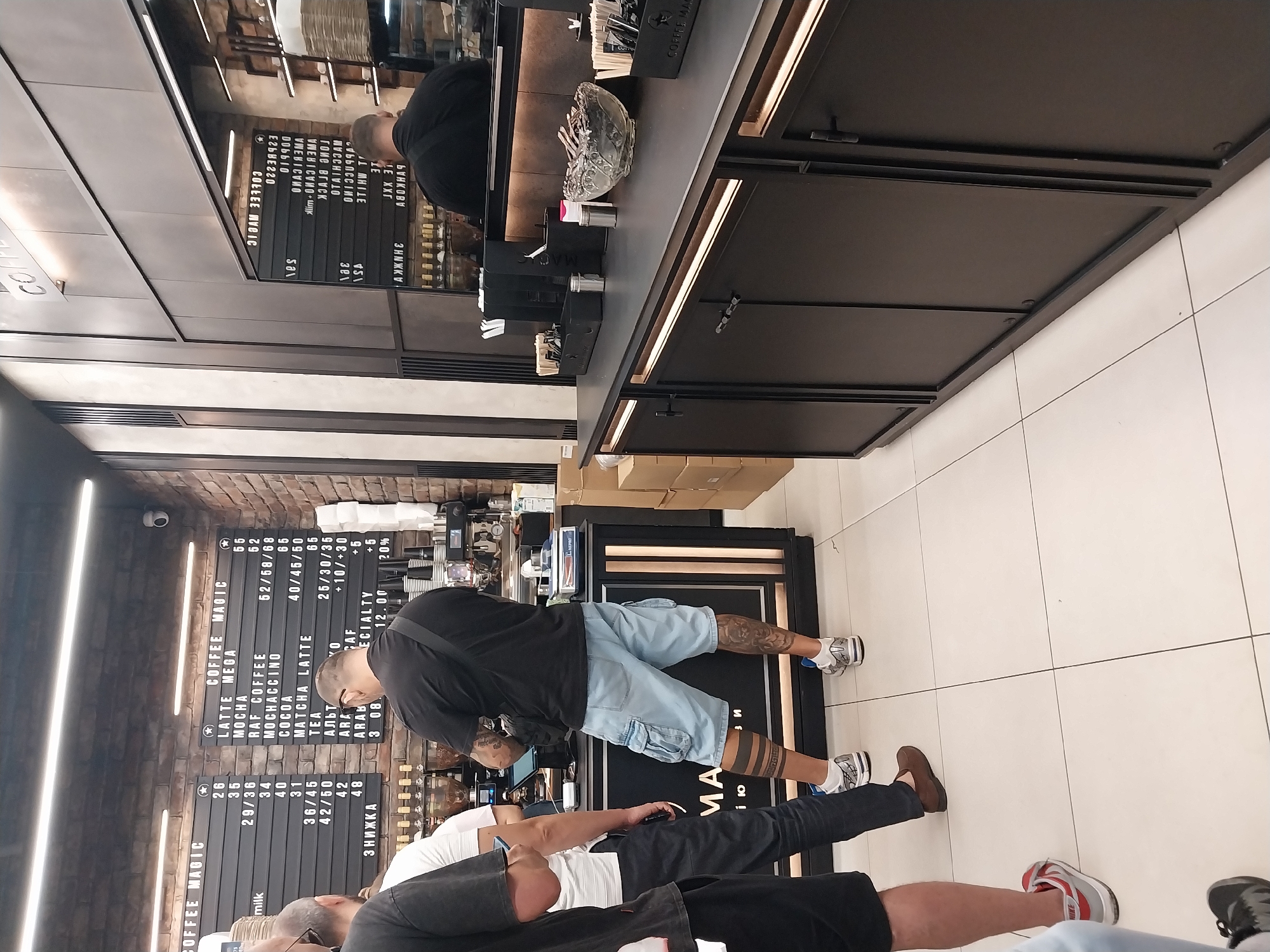 In a brightly lit, modern coffee shop that appears to be located in Dnipro, Ukraine, a customer wearing light blue cargo shorts and white athletic shoes stands at a counter, looking at a menu board. He has extensive tattoos on both of his calves. Behind him, a barista, dressed in black, is likely preparing a beverage. The menu board lists various coffee drinks and their prices. To the left, other customers are waiting in line, with one man in a black t-shirt holding a phone and another in a white t-shirt partially visible. The counter area is clean and organized, with coffee machines, grinders, and various accessories. The shop's interior features exposed brick walls and minimalist design elements, creating a contemporary atmosphere.