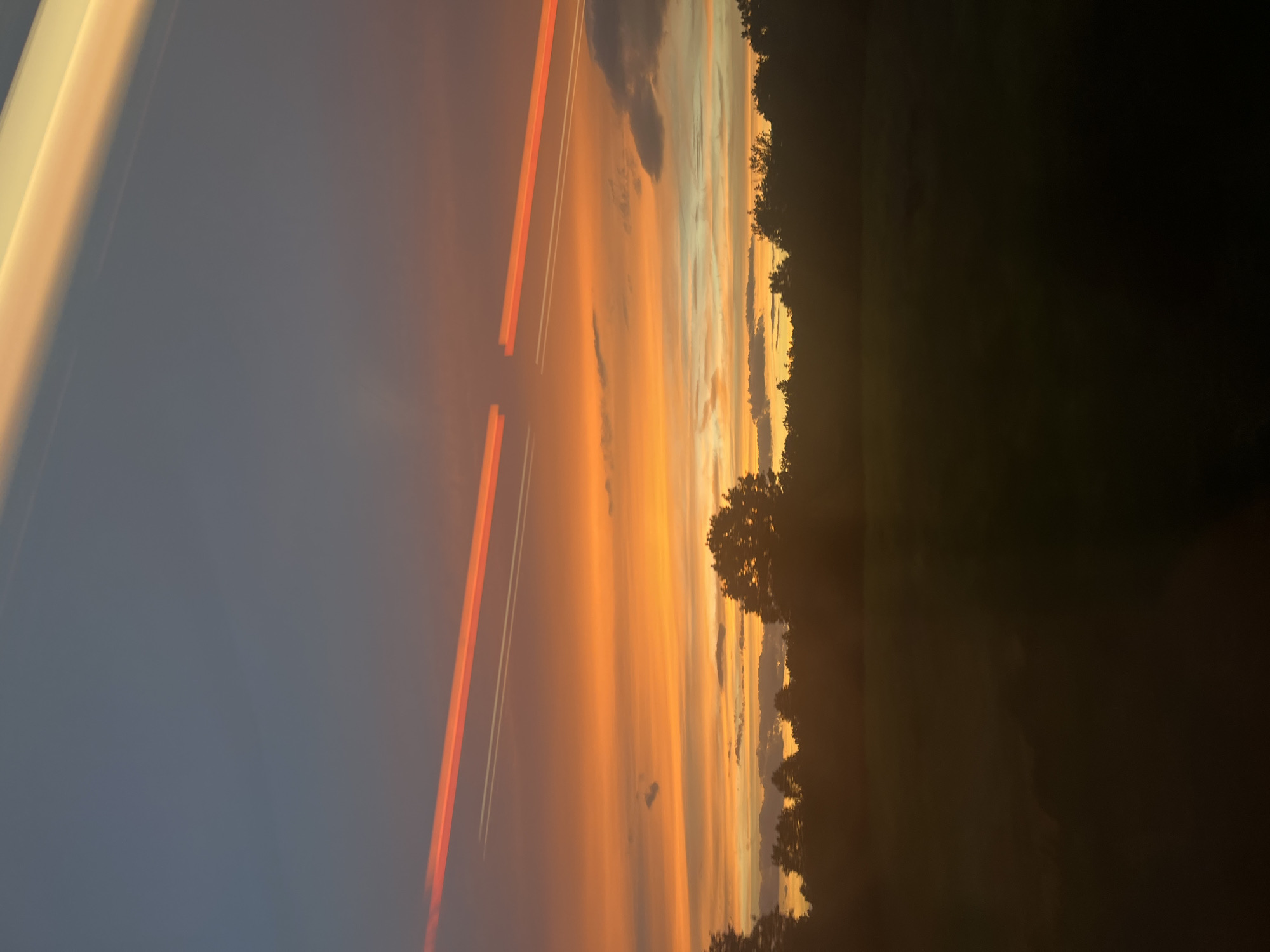 The image captures a sunset over a silhouetted treeline in Forchheim, Germany. The sky is a dramatic gradient of colors, transitioning from a deep blue at the top to vibrant oranges and yellows closer to the horizon. Two distinct contrails, one bright orange and the other white, streak across the sky from left to right, suggesting aircraft activity. Below the sky, a dark silhouette of trees forms the horizon, with a field or open space visible in the foreground, also rendered in dark, indistinct tones due to the low light. The overall scene is one of natural beauty enhanced by atmospheric phenomena and the remnants of human activity.