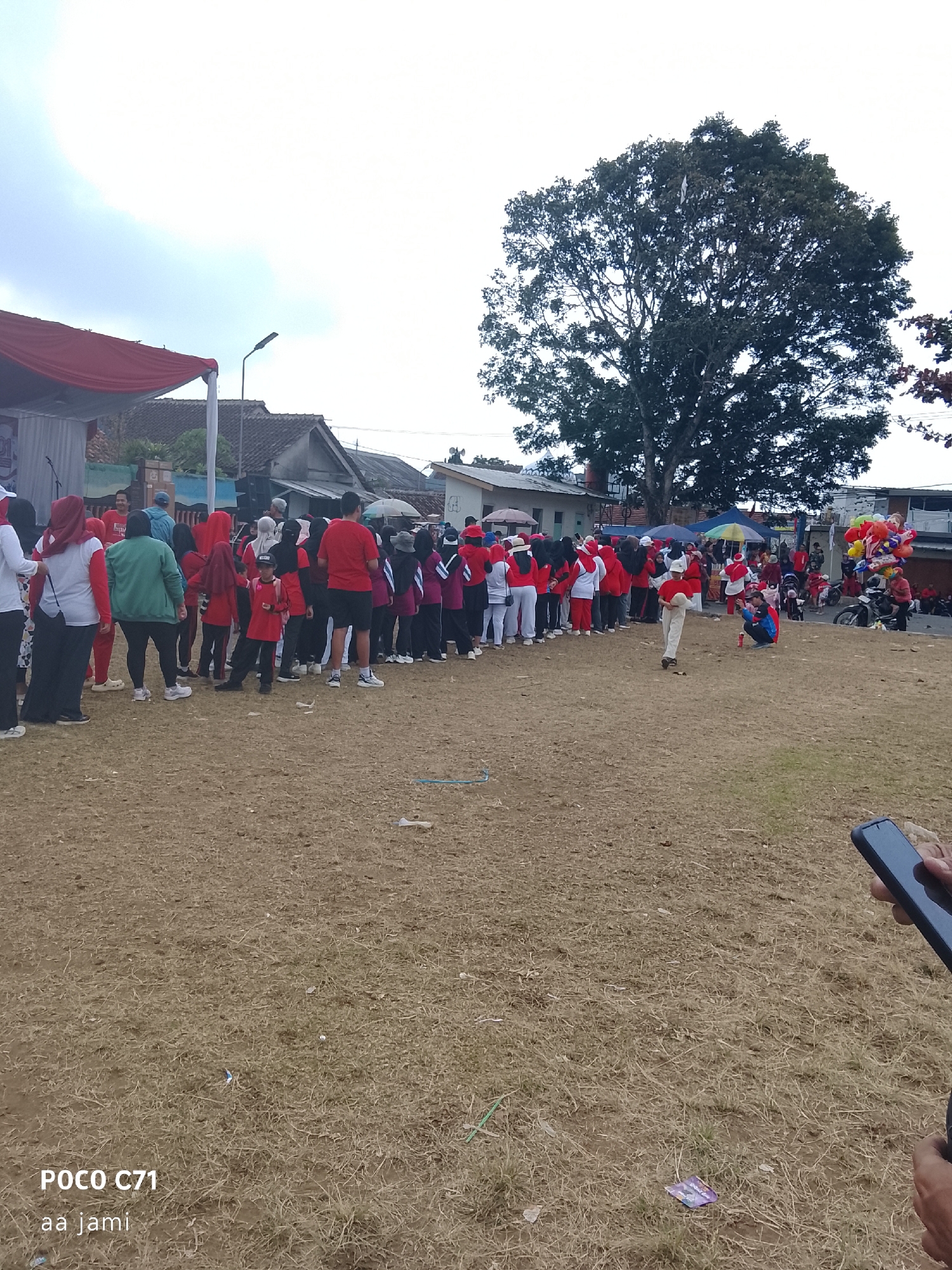 A group of people, many wearing red and white, are gathered outdoors in a dry, grassy field. The scene appears to be in Tasikmalaya, Indonesia, given the presence of local street vendors and a religious procession in the background. Some individuals are in a line, possibly participating in an event or waiting for something. There are buildings and trees in the background, suggesting an urban or semi-urban environment. Several colorful balloons are visible, and a motorcycle is parked nearby. The sky is overcast.