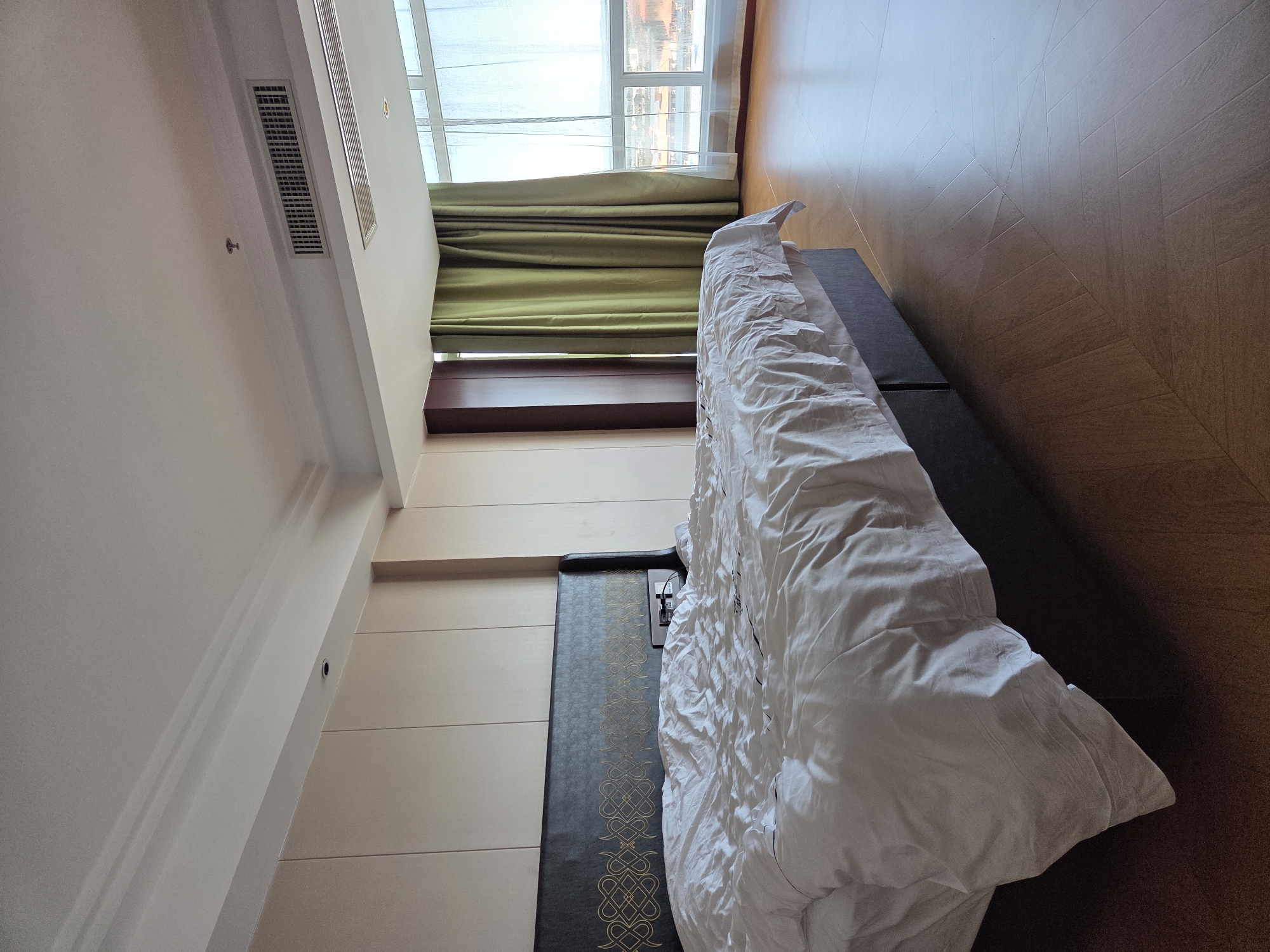 The image displays a modern bedroom or hotel room, captured from a high, slightly angled perspective looking towards a large window. A significant portion of the frame is taken by an unmade bed, covered with crumpled white bedding. Behind the bed, there's a long, dark panel featuring ornate gold, interwoven patterns, possibly a decorative headboard or wall accent. To the left of this panel, the wall is light-colored with horizontal paneling.
Natural light floods the room from a wide window at the far end, which is partially covered by sheer white curtains and olive-green drapes pulled to the side. A dark wooden shelf runs beneath the window. Beyond the window, a blurry view of an outdoor urban environment or buildings is faintly visible.
The room's ceiling and upper walls are white, incorporating multiple air conditioning vents. A small, round object, resembling a smoke detector or a tiny light fixture, is mounted on one of the walls. The floor is made of a warm-toned wood or wood-patterned material, reflecting the bright ambient light. Adjacent to the bed, on a dark, flat surface, some electronic devices or chargers are plugged into an outlet. The scene appears clean and well-lit, indicating it is daytime. No people or discernible text are present in the image. The location context is Tongjiang, China.