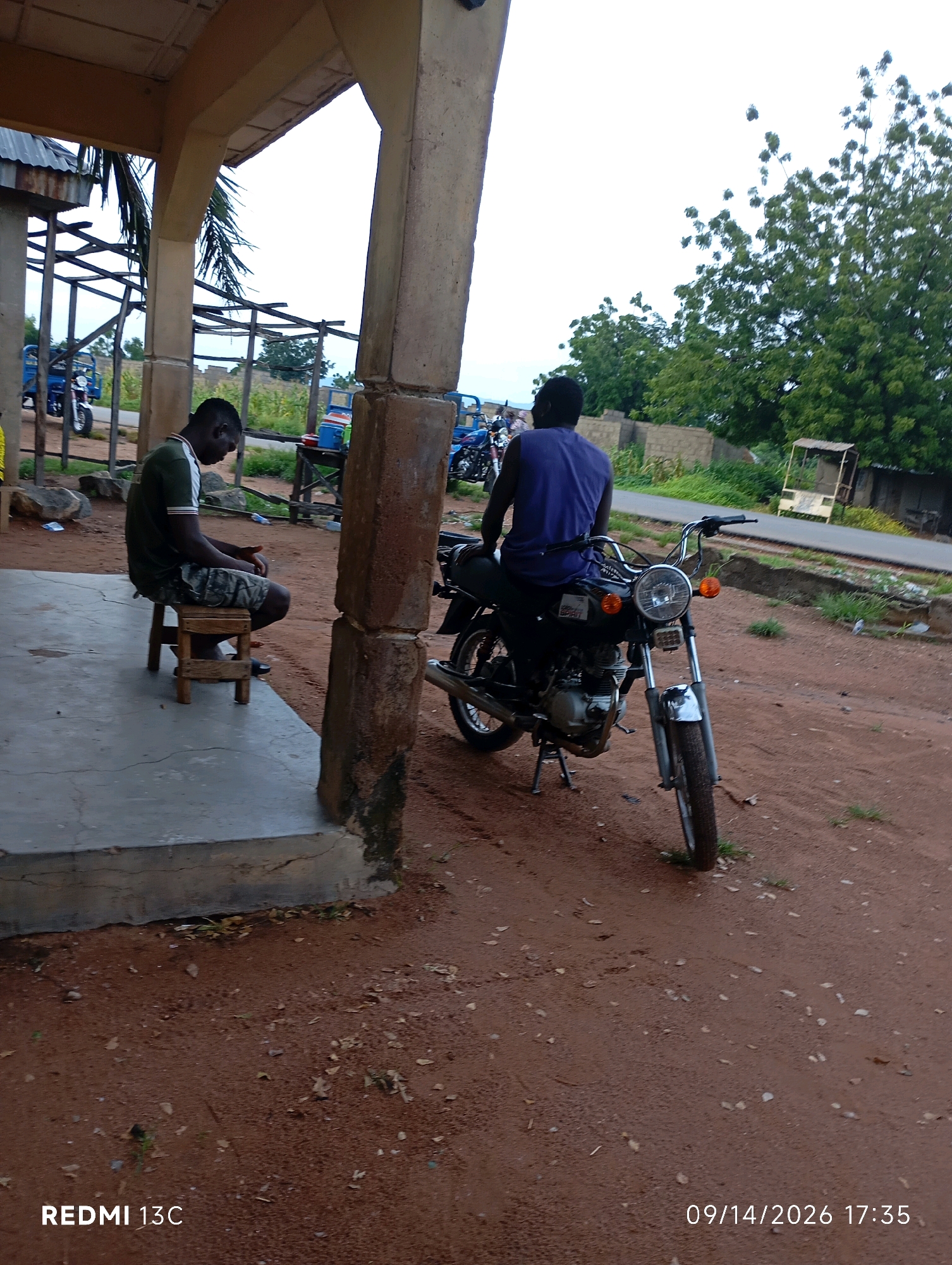 The image depicts an outdoor scene in the city of Shendam, Nigeria. In the foreground, a man is seated on a wooden stool, facing away from the camera and looking downwards. He is wearing a dark green t-shirt with white accents and camouflage shorts. Beside him, a black motorcycle is parked. Further back, another man is seated on the motorcycle, also facing away from the camera. He is wearing a blue sleeveless shirt. The environment appears to be a somewhat dusty or sandy area, with sparse vegetation and a few trees in the background. There is a covered structure with concrete pillars and a corrugated iron roof in the left portion of the image, suggesting a small shelter or an extension of a building. The ground is a reddish-brown color, consistent with the soil in the region. In the background, there are some brick structures and more trees, with a paved road visible in the distance. The sky is overcast, indicating a cloudy day. The overall impression is a casual, everyday scene in a rural or semi-urban setting.