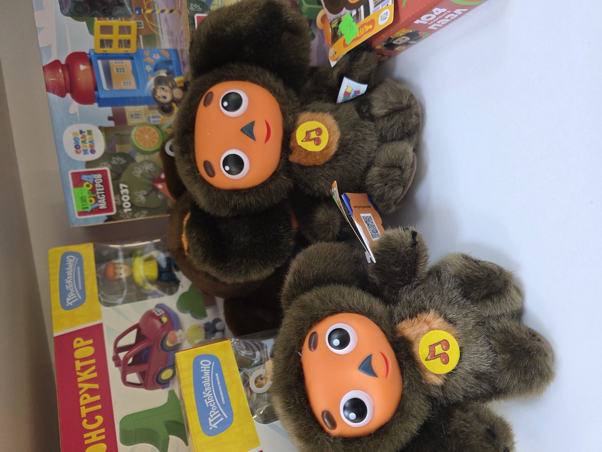 The image shows two Cheburashka plush toys lying on a white surface, with several children's construction toy sets nearby. Cheburashka, a character from Russian animation, is depicted with large round eyes, an orange face, and brown fur. Each toy has a round yellow patch with a red symbol on its chest.
The toy sets are partially visible and show colorful plastic parts. One box is labeled "CONSTRUCTOR" in Cyrillic script. Another box displays an image of a building and is labeled "SOYUZMULTFILM" and "MASTEROV 10037". Additionally, there's a box labeled "104 elements" in Russian. The text also specifies the toys are made in Russia and that the image was taken in Izhevsk, Russia.