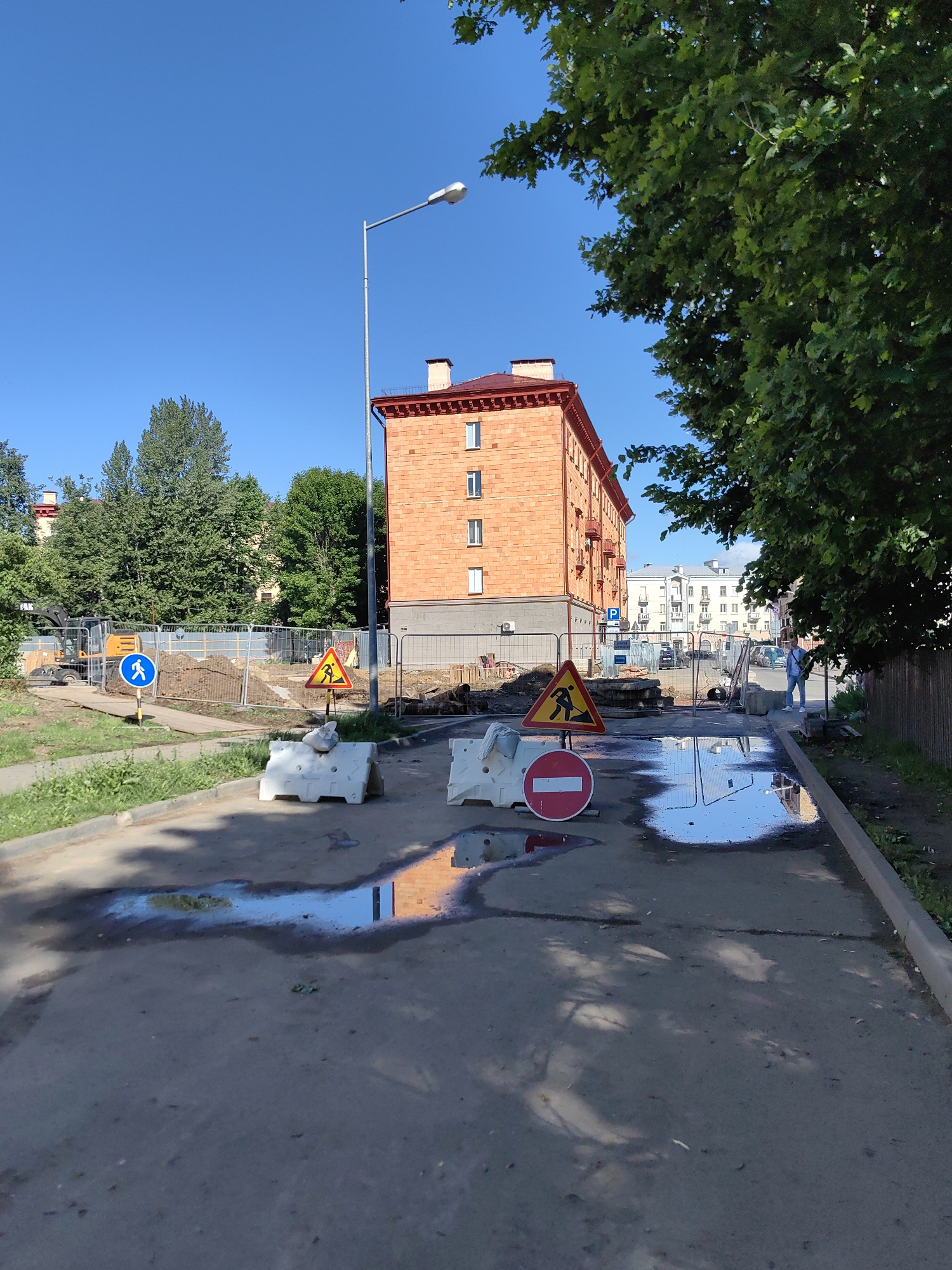 The image depicts a street scene in Minsk, Belarus, showing ongoing construction and roadwork. A large, orange-brick building with a red roof is visible in the background, flanked by trees. The foreground shows a damaged or under-construction road with several puddles reflecting the sky and surrounding buildings. Construction barriers, including white plastic traffic barriers and a metal fence, are set up to redirect traffic and pedestrians. Several traffic signs are present, including a blue circular sign with a white pedestrian silhouette indicating a pedestrian crossing, and a yellow triangular sign with a black silhouette of a person working, indicating road work ahead. A circular red and white sign, typically meaning "no entry" or "prohibited," is also visible. A person in a light blue shirt and dark pants is seen standing on the right side of the road, near the construction zone. The sky is clear and blue. The scene appears to be a typical urban environment undergoing infrastructure improvements.