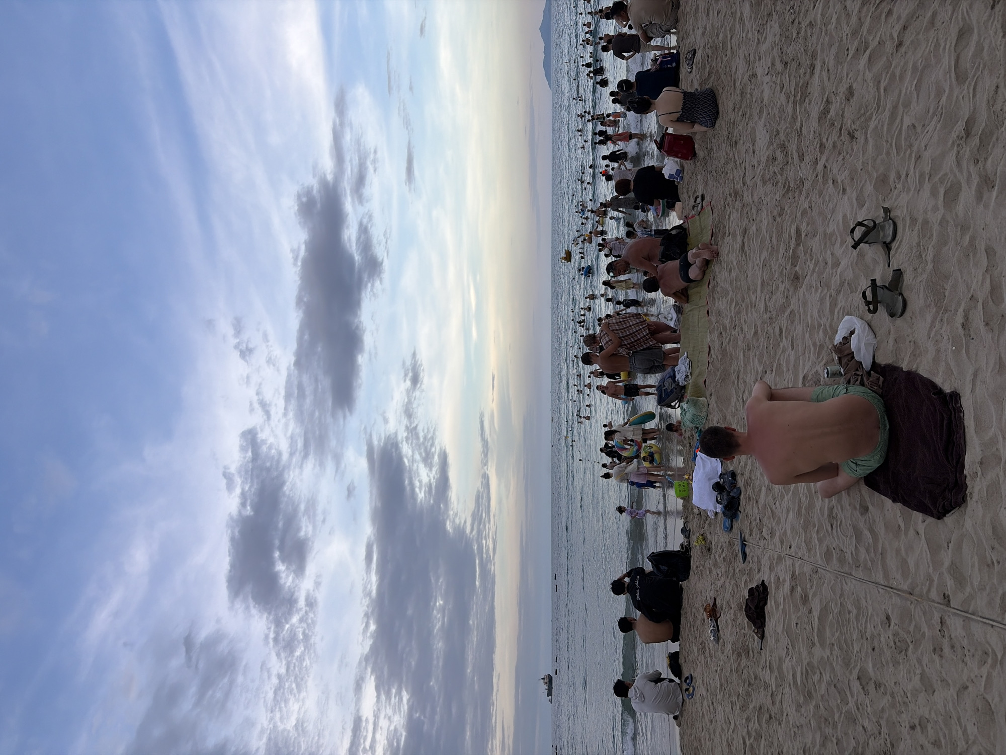 The image depicts a crowded beach scene in Đà Nẵng, Vietnam, likely during the late afternoon or early evening. The sky is partially cloudy with hues of blue, grey, and a hint of pastel pink near the horizon, suggesting the sun is setting or has recently set. The ocean is calm with small waves lapping the shore, and numerous people are visible in the water, swimming, wading, and playing. Some are standing in the shallow water, while others are further out. A small yellow boat is visible in the distance. On the beach, there is a significant gathering of people. Some are sitting or lying on towels and mats, while others are standing or walking. The sand is light brown and textured, with footprints and scattered debris such as towels, bags, and sandals. In the foreground, a man is sitting on a towel facing away from the camera, wearing green shorts. Further back, several groups of people are gathered, some appearing to be families or friends. A large cargo ship is visible on the horizon to the left. The overall atmosphere appears to be one of leisure and recreation.