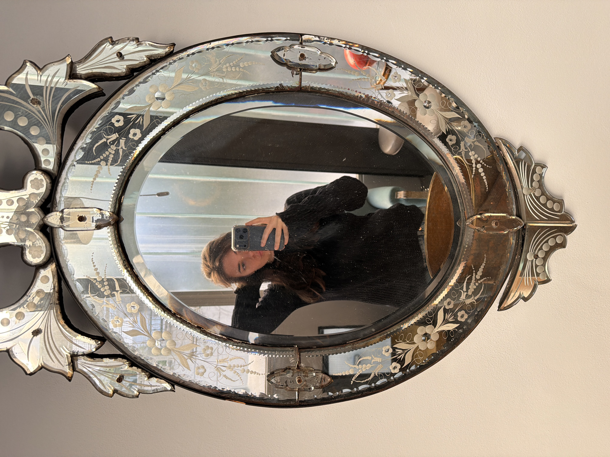 The image displays a selfie captured through the reflection of an ornate, vintage mirror. The central reflective area of the mirror is oval, framed by an elaborate border of etched and beveled glass panels. These panels are intricately decorated with floral and foliate designs, including delicate flowers, leaves, vines, and dotted patterns, giving the mirror a distinct antique or Venetian style.
The mirror's frame exhibits signs of age, with visible tarnishing or oxidation on the silvering, appearing as darker, almost bronze-toned areas, and some minor imperfections on the glass edges. Decorative metal fasteners secure the different glass components of the frame.
In the reflection, a young woman with long, dark brown hair is seen holding a smartphone (an iPhone model with a prominent camera array) to take the picture. She is wearing a dark, possibly black, ribbed long-sleeved top, and her head is slightly tilted towards the camera.
The reflection also offers glimpses of an interior space, including what appears to be a light source, possibly a window, and indistinct household items like a counter or table, suggesting a domestic setting. The wall behind the mirror is a plain, light, off-white color. The scene is well-lit, indicating it is daytime. No discernible text is visible on the mirror, the woman's attire, or any reflected objects. The context for the image is Buenos Aires, Argentina.