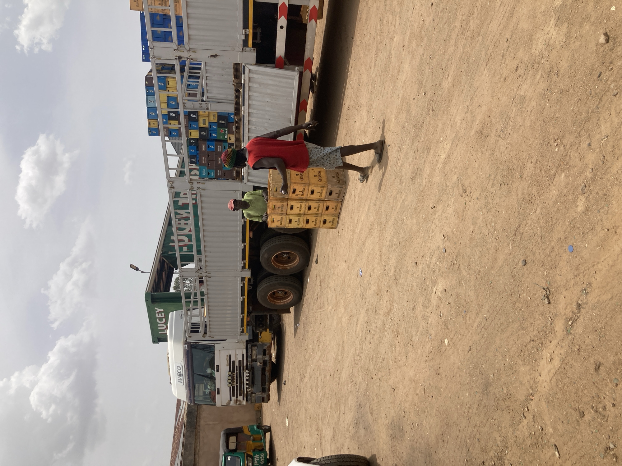 The image depicts a scene near a parked truck in Bukuru, Nigeria. The truck, marked with "LUCEY" and "IVECO," is loaded with crates of Guinness beer. Two men are unloading the crates; one is carrying a stack of them. The ground is dirt and dusty. A three-wheeled vehicle is parked nearby. The sky is partly cloudy.