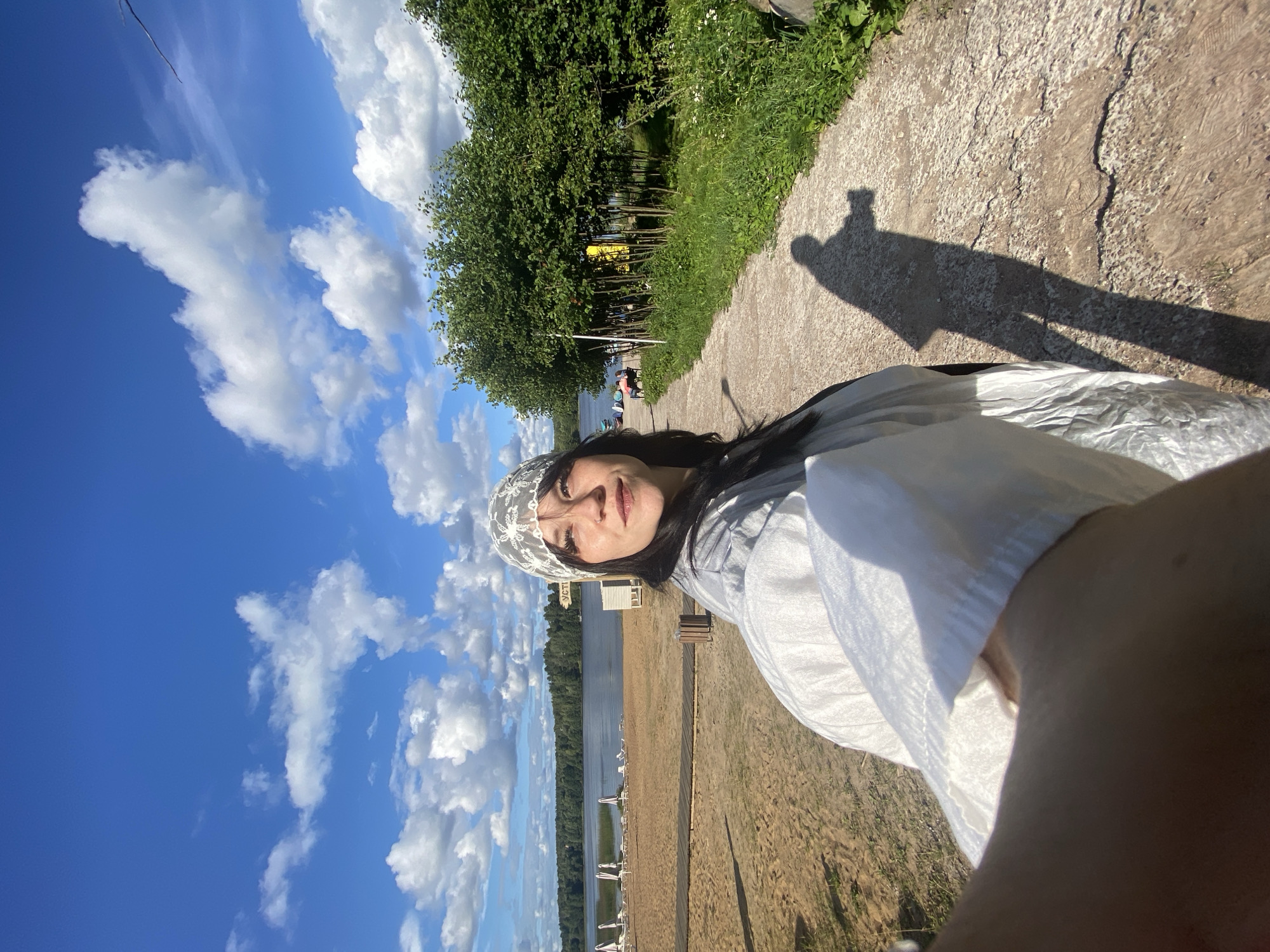 A selfie is captured by a woman standing on a path next to a sandy beach. She is wearing a white bandana and a light-colored jacket. The background features a lake with lounge chairs on the beach, a line of trees, and a bright blue sky with white clouds. A shadow of the woman is cast on the path in front of her. The scene is set in Krasnoflotskoe, Russia.
