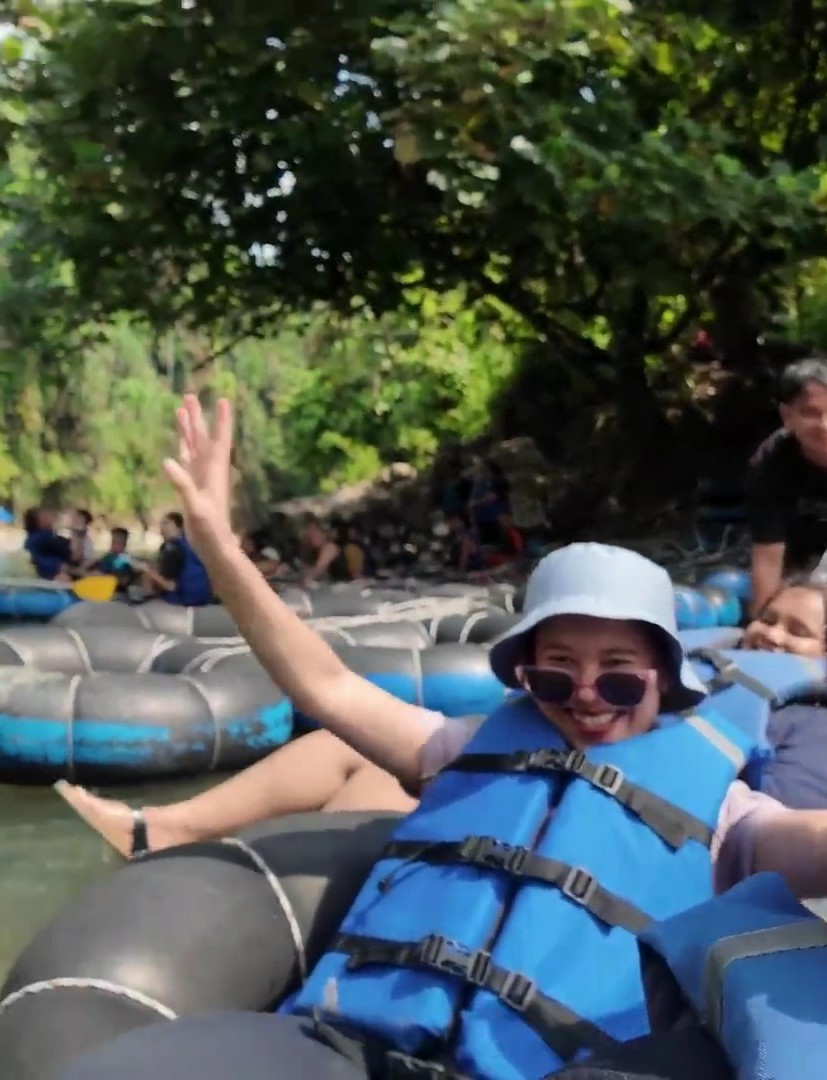 In the foreground, a young woman wearing a light blue bucket hat, sunglasses, and a bright blue life jacket is tubing down a river. She is smiling and raising her right hand, her left leg extended. She is in a black inflatable tube, and another person is partially visible behind her in a similar tube. In the mid-ground, several other inflatable tubes, some with people in them, are also on the river. The background shows lush green trees and foliage lining the riverbanks, with a rocky slope to the left. The water is calm and appears shallow. The scene suggests an outdoor recreational activity in a natural, wooded environment.
