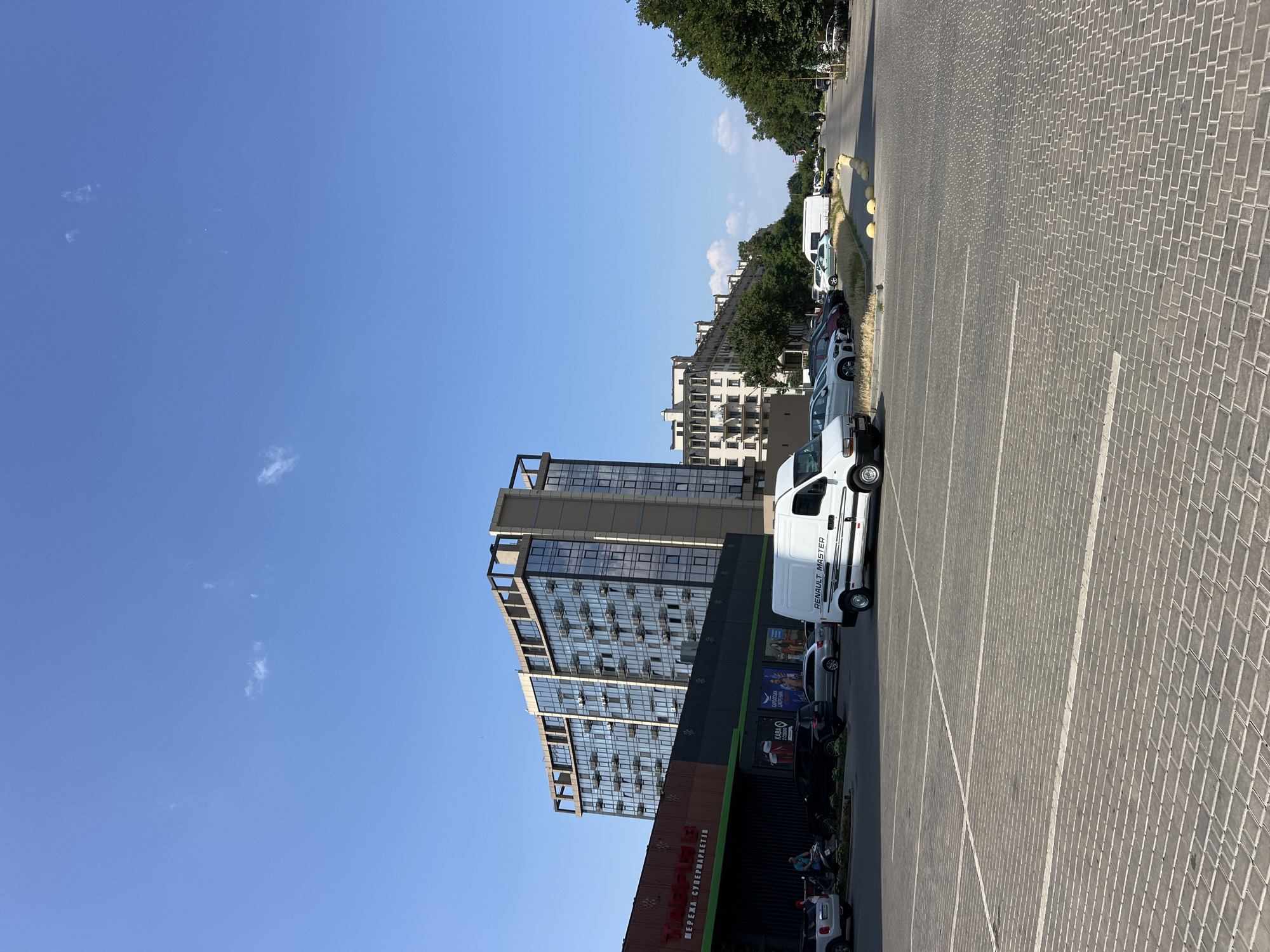 The image depicts a scene in Fontanka, Ukraine, on a clear, sunny day. In the foreground, a paved parking lot is visible, marked with white lines. A white Renault Master van is parked prominently in the lot. To the left, a supermarket with a dark facade and red lettering is situated. Behind the supermarket, a modern, multi-story building with a glass facade and balconies dominates the mid-ground. Further down the street, a row of apartment buildings with similar architectural styles lines the road, bordered by a line of lush green trees. Several cars are parked along the street and in the supermarket parking lot. The sky is a bright, clear blue with a few scattered white clouds.