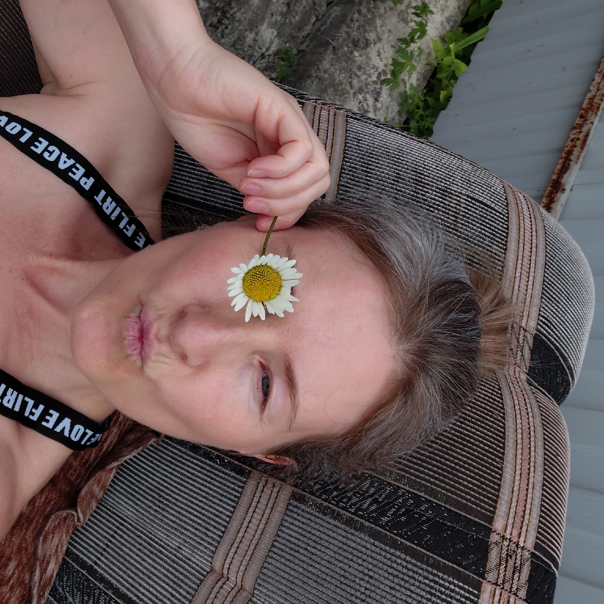 The image depicts a woman lying on a striped chaise lounge outdoors, with a daisy held up to her left eye. She is wearing a black bra with white lettering that reads "FLIRT PEACE LOVE". Her hair is pulled back, and she is puckering her lips. The background shows a grey metal fence and some greenery. The scene is likely a backyard or patio. There is no information in the image that allows for the identification of the city of Novosibirsk, Russia.
