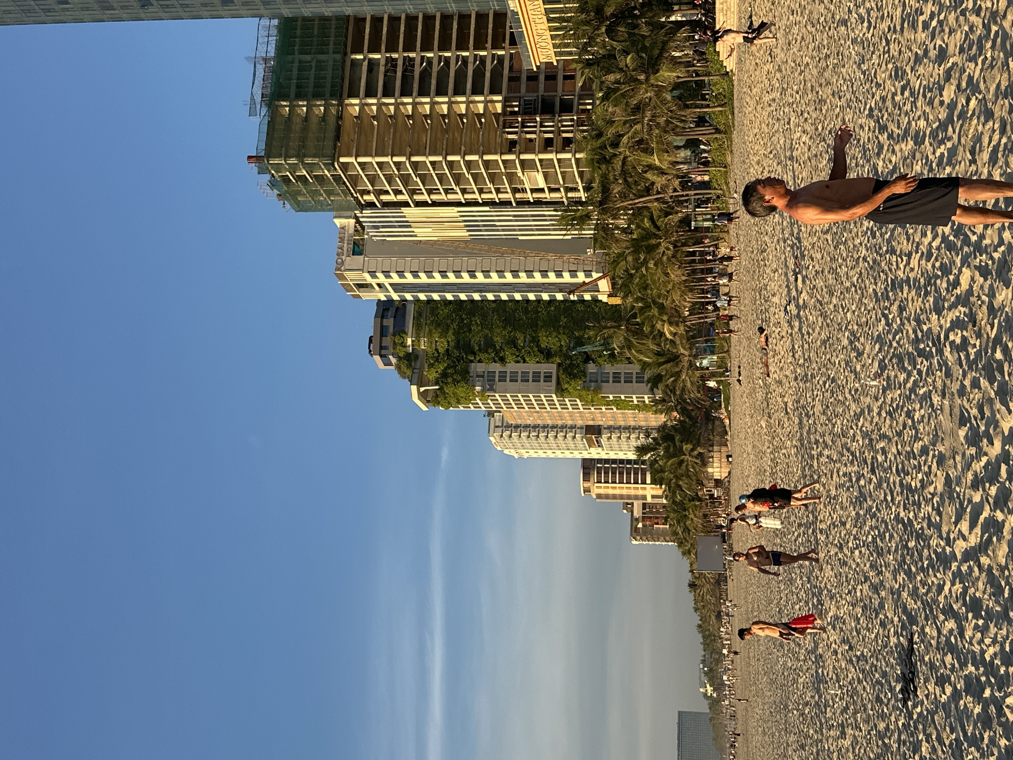 This is a photograph taken on a beach in Đà Nẵng, Vietnam, likely at sunset given the lighting. In the foreground, a man with dark hair is facing right, wearing black shorts, standing on a sandy beach. Behind him, other people are visible on the beach, some walking and some standing.
In the background, a line of buildings stretches along the coastline. Several of these appear to be high-rise hotels or residential buildings, with some in various stages of construction, identifiable by scaffolding and exposed concrete. Palm trees are planted in front of the buildings, creating a tropical landscape. The sky is a clear, vibrant blue with some wispy clouds visible. The overall impression is a busy coastal urban environment at the end of the day.