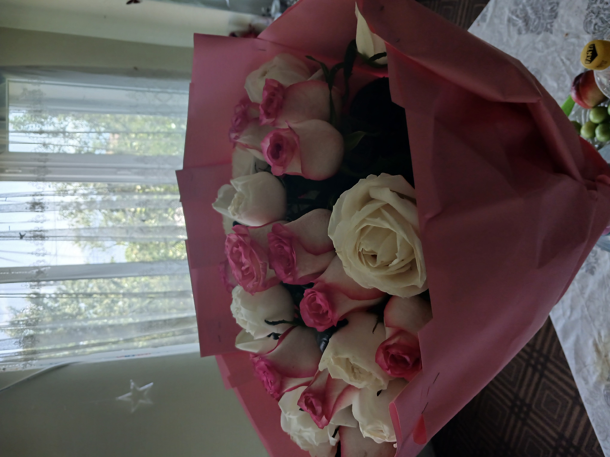 A bouquet of white and pink roses is prominently displayed in the foreground of the image. The flowers are wrapped in pink paper. Behind the bouquet, a window covered by sheer, white lace curtains reveals a blurred view of green foliage, suggesting an outdoor environment. The room appears to be lit by natural light streaming through the window. To the left of the window, a white, star-shaped ornament hangs from a string. The table in the lower portion of the image is partially visible, with a patterned tablecloth. A yellow bottle cap with the word "FANTA" is visible on the right side of the table, along with some green fruits and a red fruit. The scene is indoors, in what appears to be a domestic setting, likely in Telavi, Georgia, given the context of previous interactions.