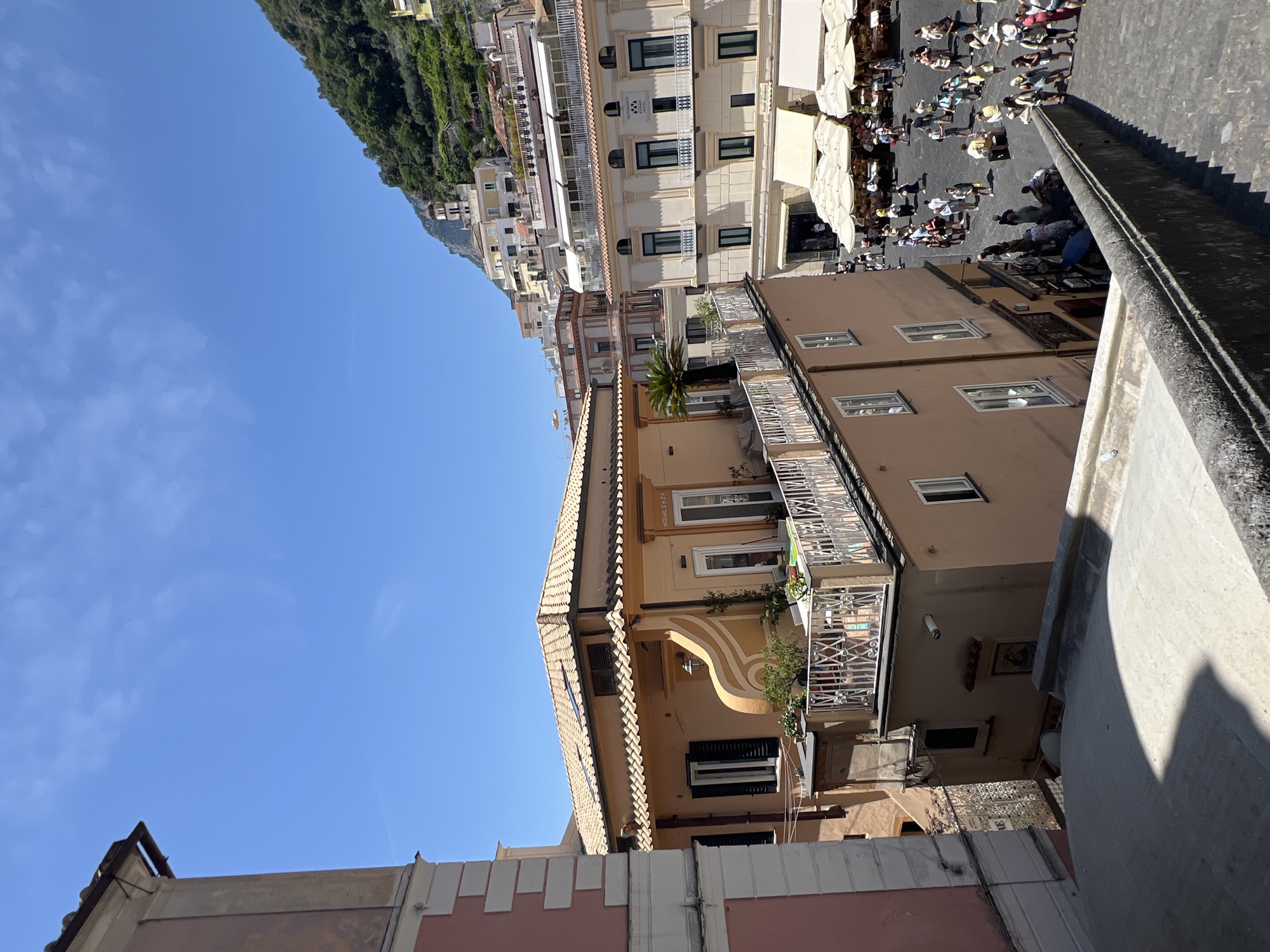 This is a photograph taken on a sunny day in Pogerola, Italy. The scene is a bustling street with many people walking around. Buildings line the street, some of which appear to be shops or restaurants with outdoor seating. The architecture suggests a Mediterranean setting, with stucco buildings and tiled roofs. A steep, tree-covered hillside rises behind the buildings, adding to the dramatic landscape. The time of day appears to be midday, given the bright sunlight and the shadows cast by the buildings and umbrellas. The atmosphere seems lively and energetic, with many people enjoying the pleasant weather and the vibrant street life. There is no visible text in the image.