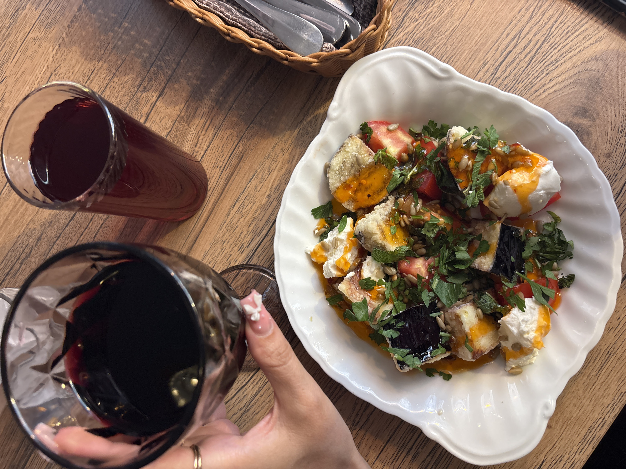 An overhead, close-up shot shows a person's left hand, adorned with a ring and decorative fingernails, holding a glass of dark red wine. Adjacent to it is a tall glass filled with a similar red liquid. A white, scalloped plate, overflowing with a colorful salad composed of diced tomatoes, chunks of white cheese, dark purple eggplant slices, and fresh herbs, sits on a wooden table. Drizzled over the salad is an orange dressing, and scattered throughout are sunflower seeds. To the right of the plate, a woven basket holds cutlery. The setting appears to be a restaurant.