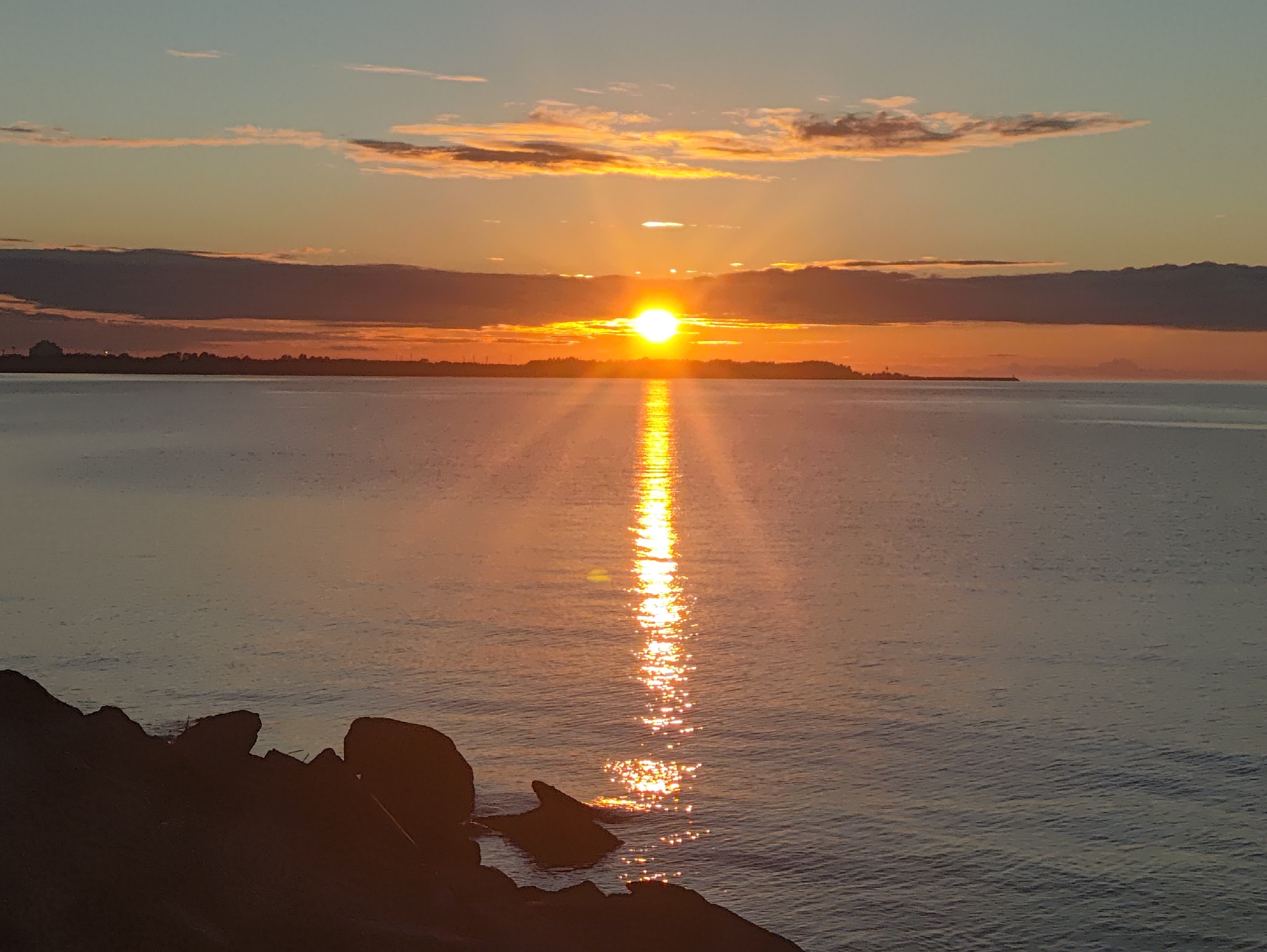 This image depicts a picturesque sunset over the Baltic Sea, likely viewed from the coast of Tallinn, Estonia. The sun, a bright orb, is low on the horizon, casting a brilliant golden path across the calm water. The sky is a gradient of soft blues and oranges, with scattered clouds tinged with sunset hues. In the distance, a dark, silhouetted landmass is visible, representing the far shore or a peninsula within Tallinn. The foreground features a dark, rocky shoreline, providing a contrasting texture to the smooth water and sky. The overall atmosphere is serene and tranquil, highlighting the natural beauty of the coastal environment at dusk.