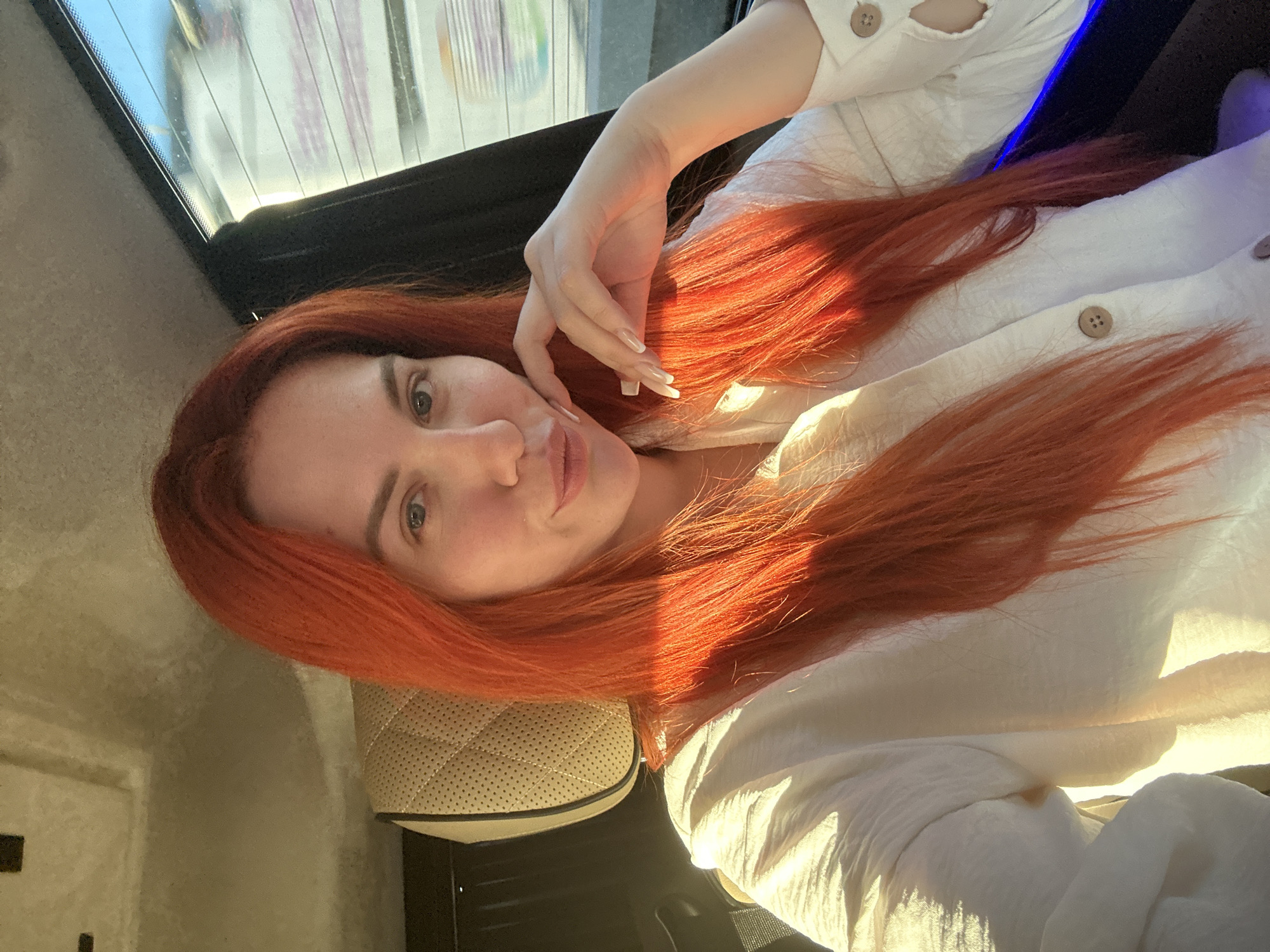 A woman with vibrant red hair sits in the passenger seat of a car, facing the camera. The interior of the car appears to be luxurious, with quilted leather headrests. Sunlight streams through the window, casting a warm glow on her face and white button-up shirt. Her left hand is raised, with manicured nails resting against her chin. In the background, through the car window, a blurry, out-of-focus view of the outside environment suggests movement, possibly on a street in Kâğıthane, Turkey. The car's interior lighting, with hints of blue, is also visible.