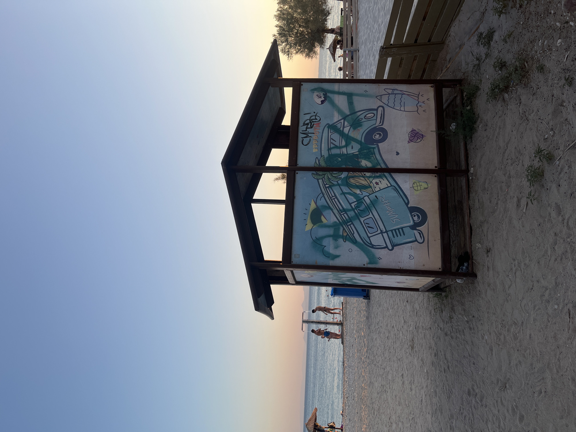 This is a photograph of a beach in Paleo Faliro, Greece, during sunset. The sky is a gradient of blue and orange, transitioning into a soft pink horizon. In the foreground, a wooden structure with a covered roof stands on the sand. The front panel of this structure is adorned with graffiti, featuring a cartoonish blue camper van with the word "SUMMER" on it, along with other drawings of surfboards, a basketball, and a drink. To the left of this structure, a blue trash bin is visible.
Further into the background, on the sandy beach, a shower stall is set up, with three individuals near it. Two men appear to be showering, while a third, smaller figure stands nearby. Beyond the shower, the sea stretches out to the horizon, meeting the sky. A beach umbrella and lounge chairs are visible on the right side of the image, where more people are seated. A tree with a rounded canopy stands to the right of the structure. The overall scene depicts a relaxed atmosphere on a beach as the day comes to an end.