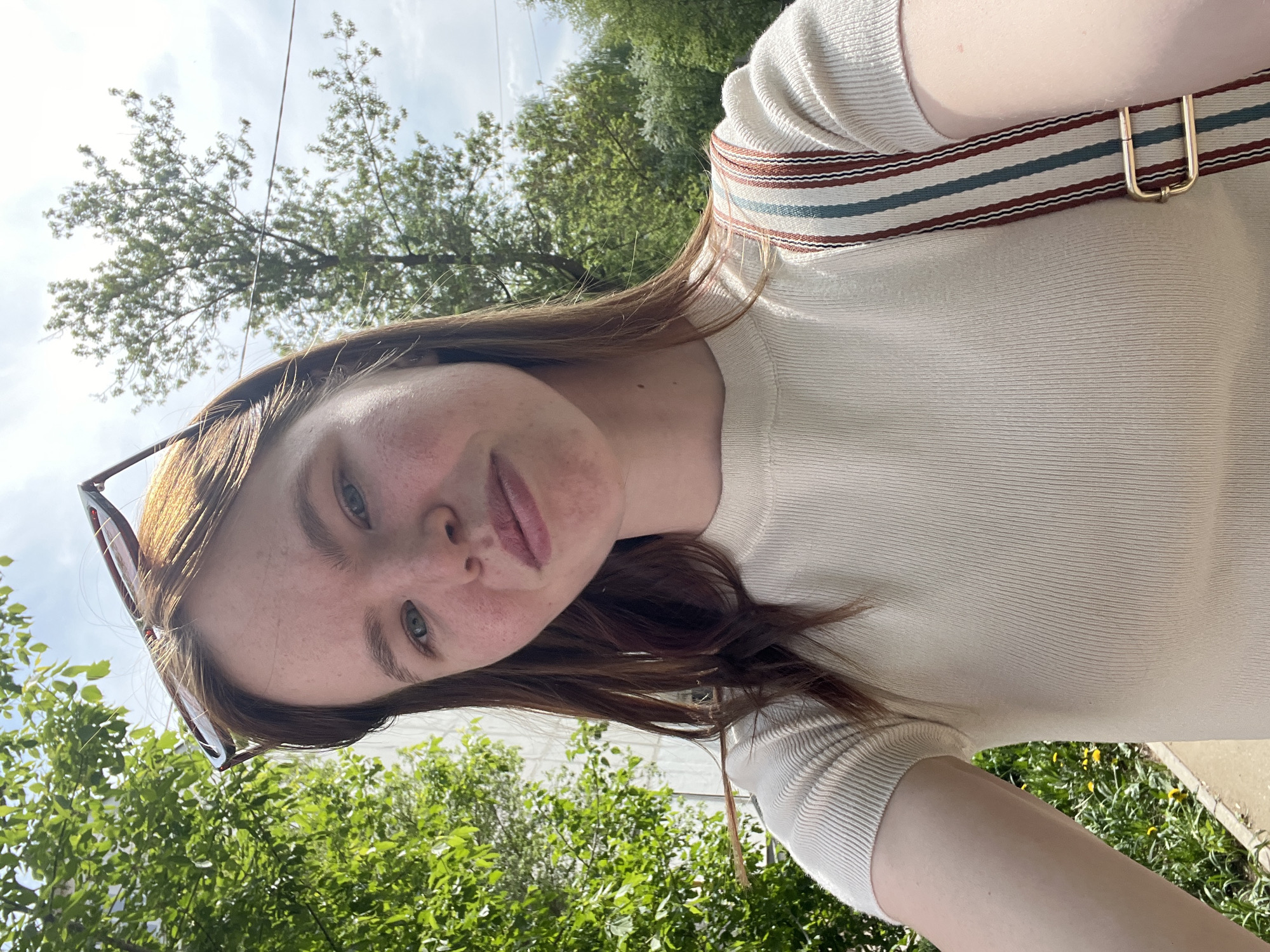A selfie shows a fair-skinned person with freckles, reddish-brown hair, and glasses propped on their head. They are wearing a light-colored, ribbed top with a striped strap slung across their shoulder. The background consists of green trees and a sky with light clouds. Some green grass and dandelions are visible in the lower right. The location is not discernible from the image.