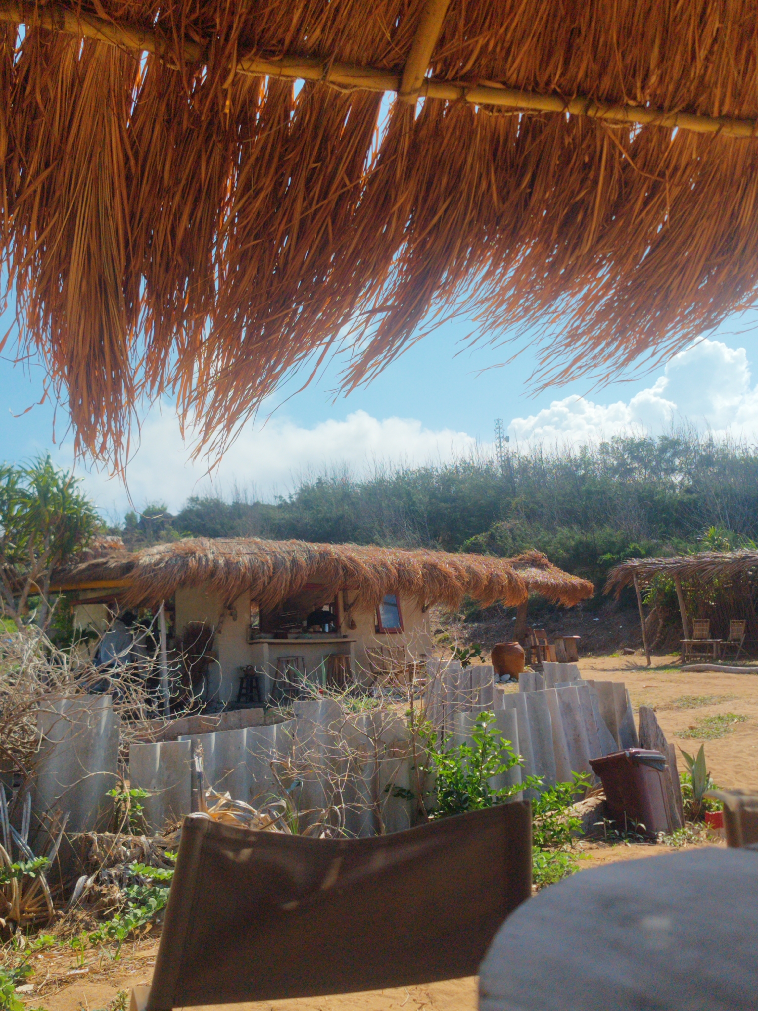The image shows an outdoor scene in Phú Quý Special Administrative Region, Vietnam. In the foreground, a partially visible chair and table suggest a seating area. Behind this, a low, corrugated fence separates the foreground from a building with a thatched roof. The building appears to be a small, open-air structure, possibly a bar or cafe, with a counter and some seating visible inside. Adjacent to the building is another thatched-roof structure, seemingly an open shelter with wooden chairs and a table. The background features a hill covered in vegetation. A blue sky with scattered clouds is visible above the thatched roofs and between the foreground foliage. The scene appears to be a casual outdoor setting, likely a tourist or leisure area.
