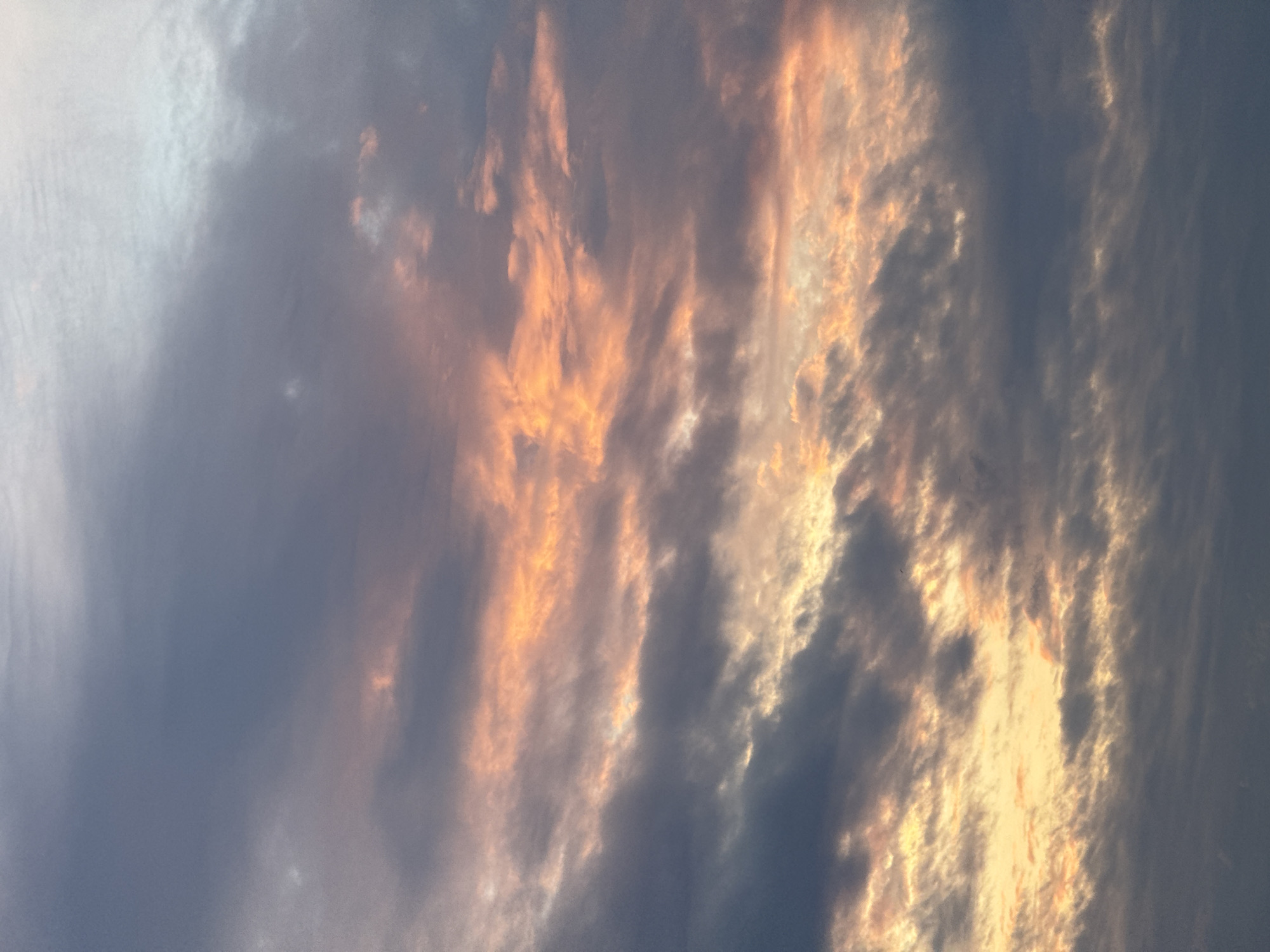 The image displays a sky filled with dramatic clouds during what appears to be sunset or sunrise. The clouds are a mixture of dark grays and lighter shades, with streaks of warm orange and pink hues illuminating them from behind. These colors suggest the sun is low on the horizon, casting its light through the atmospheric particles. The cloud formations are varied, with some appearing wispy and ethereal, while others are more dense and layered, creating a sense of depth and texture. There is no discernible geographical location within a city visible, nor are there any people, subjects, objects, or actions depicted. The scene is solely focused on the atmospheric phenomenon of the sky.