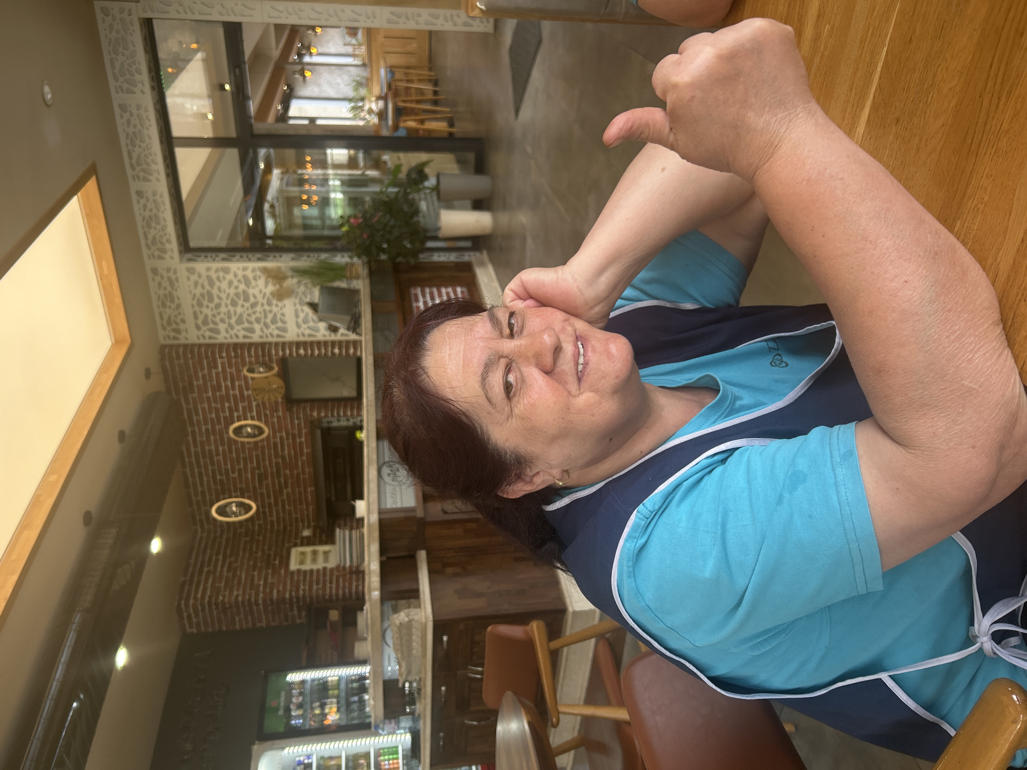 A middle-aged woman with dark brown hair smiles and gives a thumbs-up. She wears a light blue t-shirt with a dark blue vest and white trim. The image appears to be taken inside a restaurant or cafe, possibly in Kazanlak, Bulgaria. Behind her, a brick wall is visible, decorated with lights. Through a large window, one can see greenery and tables. A refrigerator is visible to the left. The seating appears to be leather chairs.
